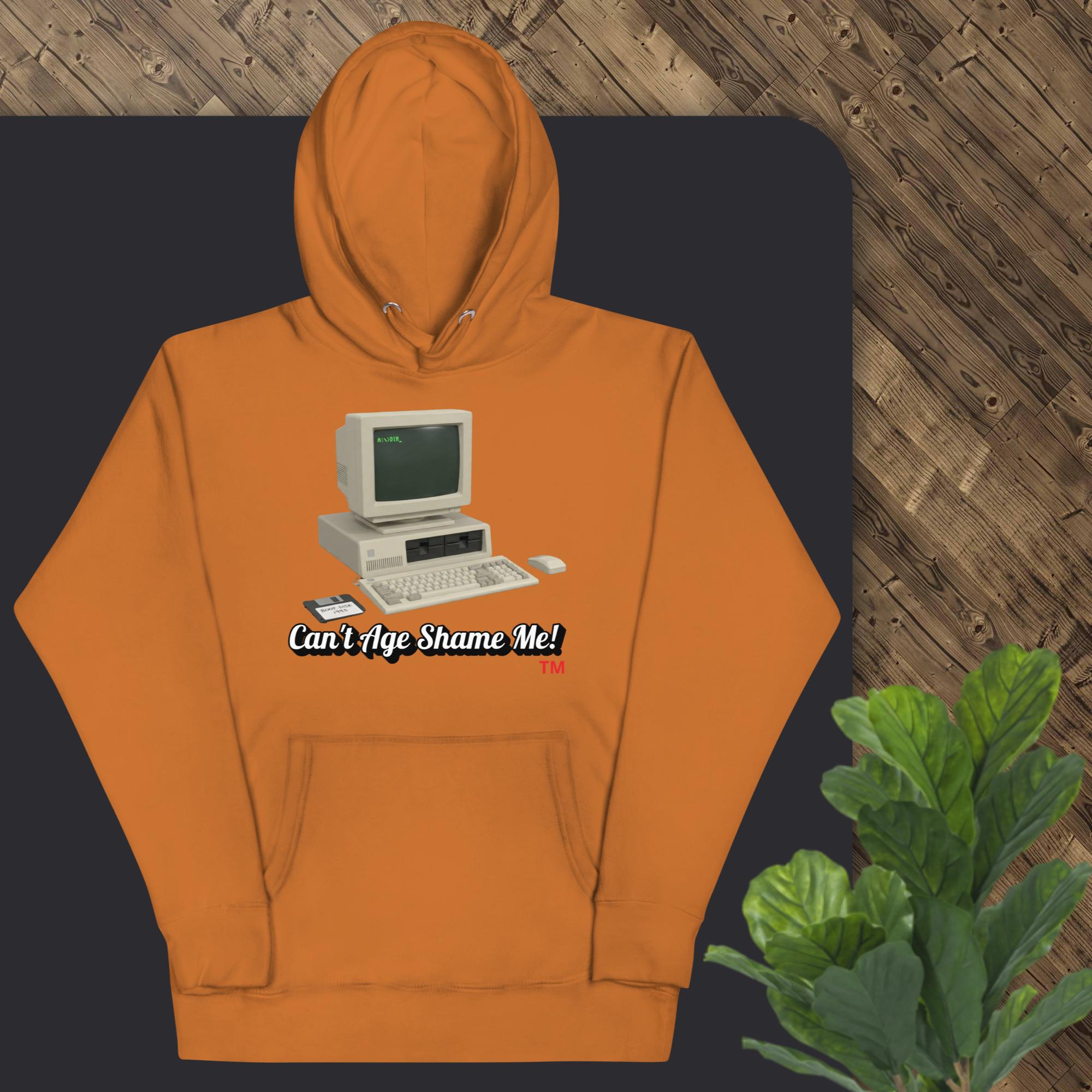 adobe  hoodie with vintage computer graphic and text 'Can't Age Shame Me!' on a dark surface.