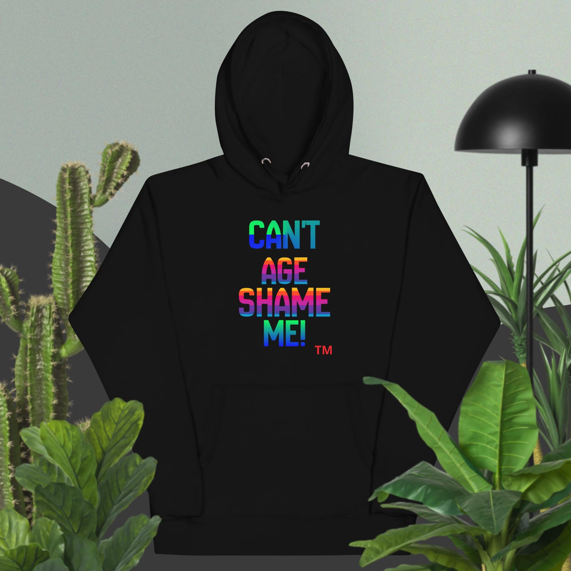 Black hoodie with colorful text 'CAN'T AGE SHAME ME!' in a room with plants and a lamp.