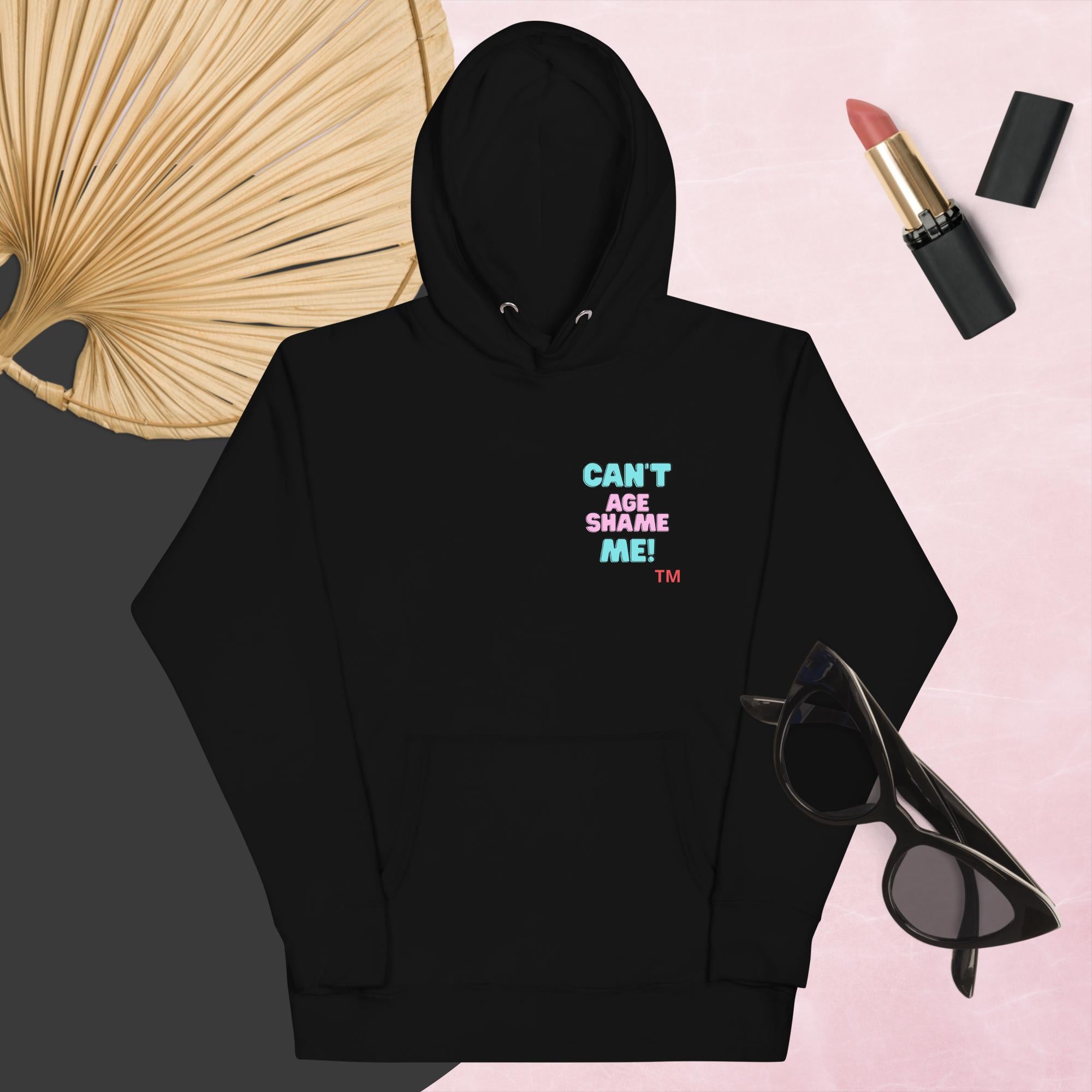 Black hoodie with text, 'Can't Age Shame Me' with sunglasses, lipstick, and a decorative leaf on a pink background