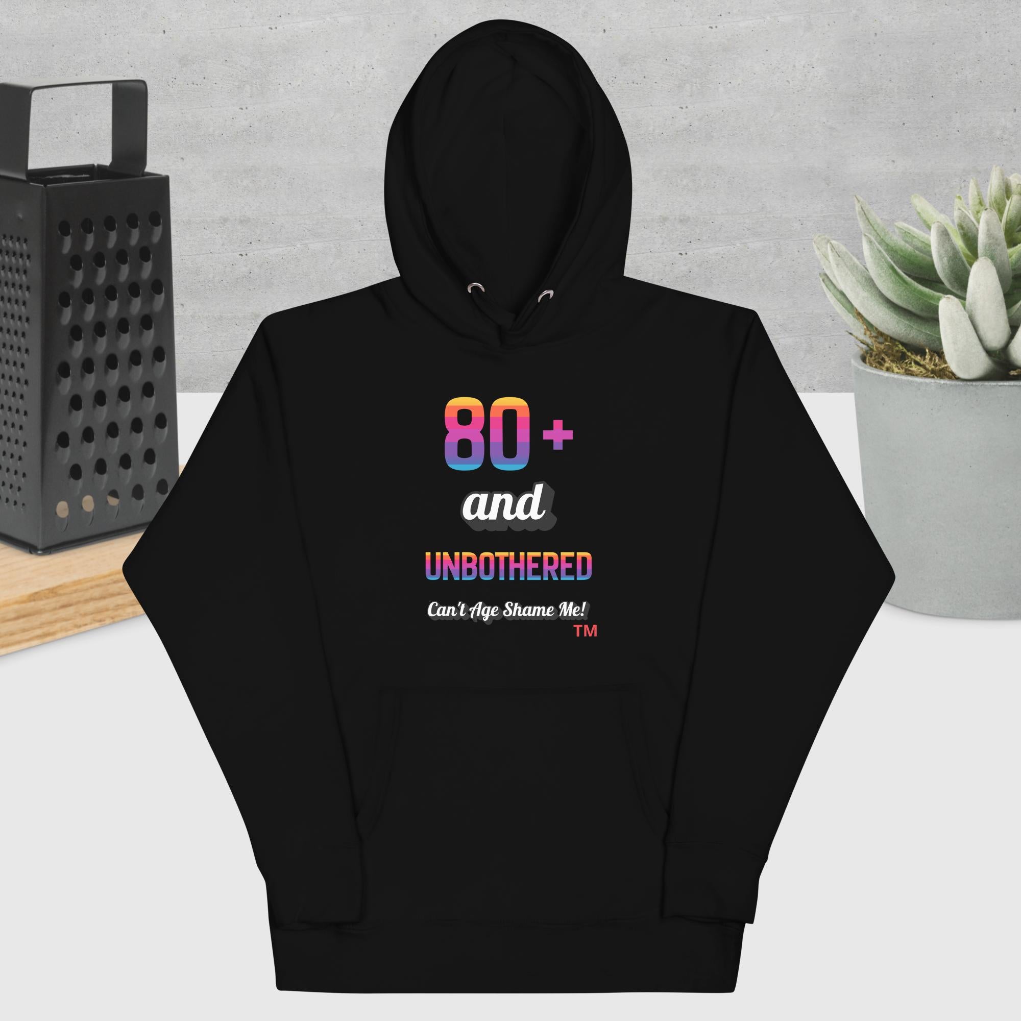 Black hoodie with colorful text design on a gray surface with a plant and box in the background