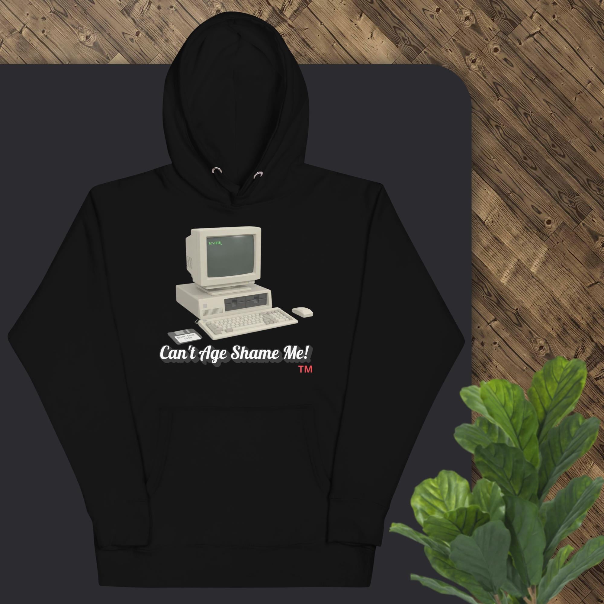 Black hoodie with vintage computer graphic and text 'Can't Age Shame Me!' on a dark surface.