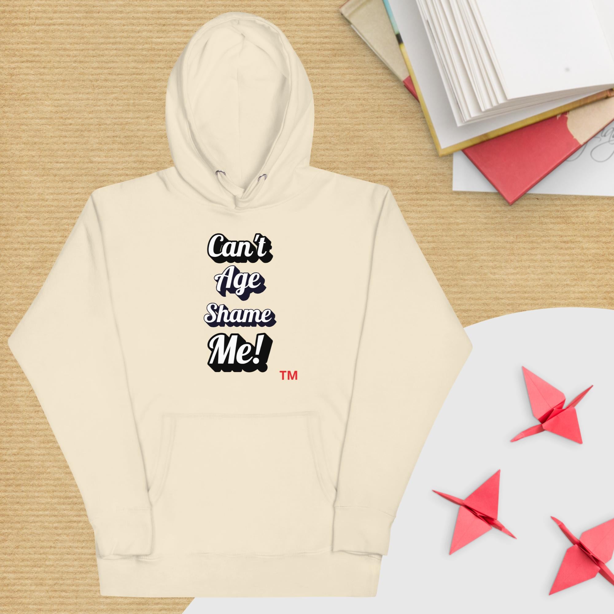 Beige hoodie with 'Can't Age Shame Me!' text on a wooden surface with books and paper cranes.
