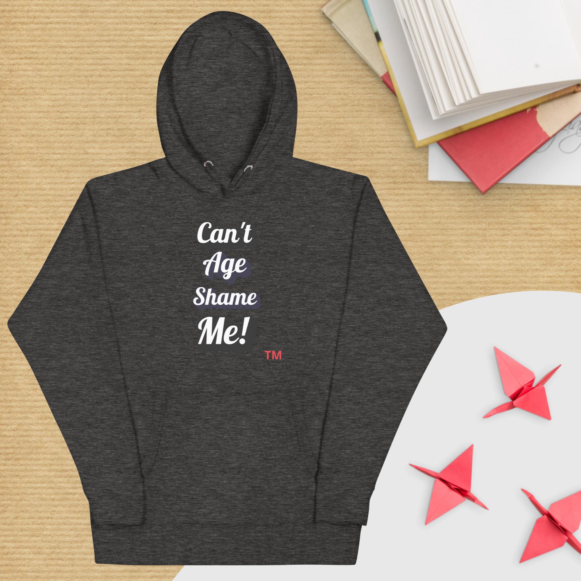 Dark gray hoodie with 'Can't Age Shame Me!' text on a wooden surface with books and paper cranes.