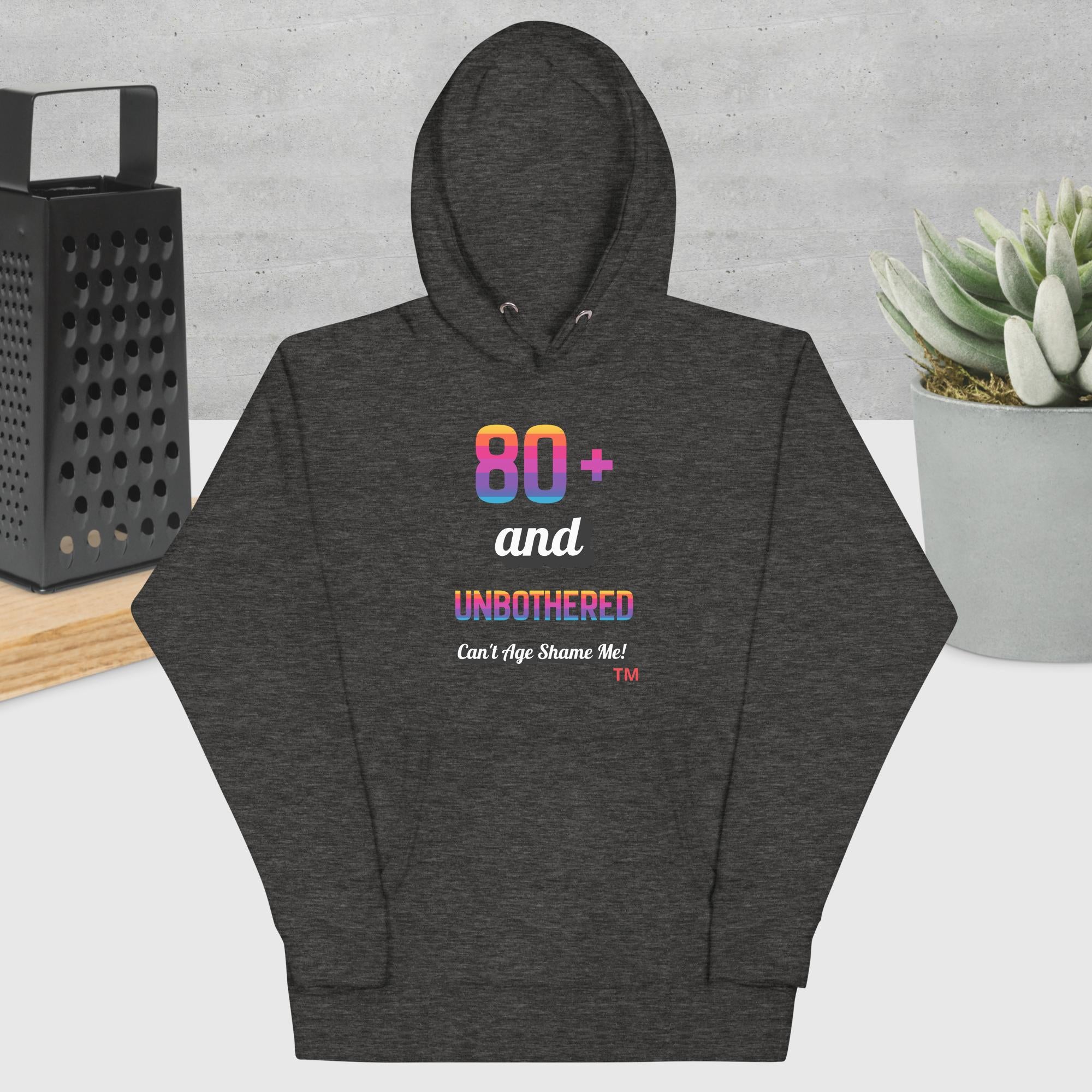 Dark gray hoodie with colorful text design on a light surface with a plant and black box in the background