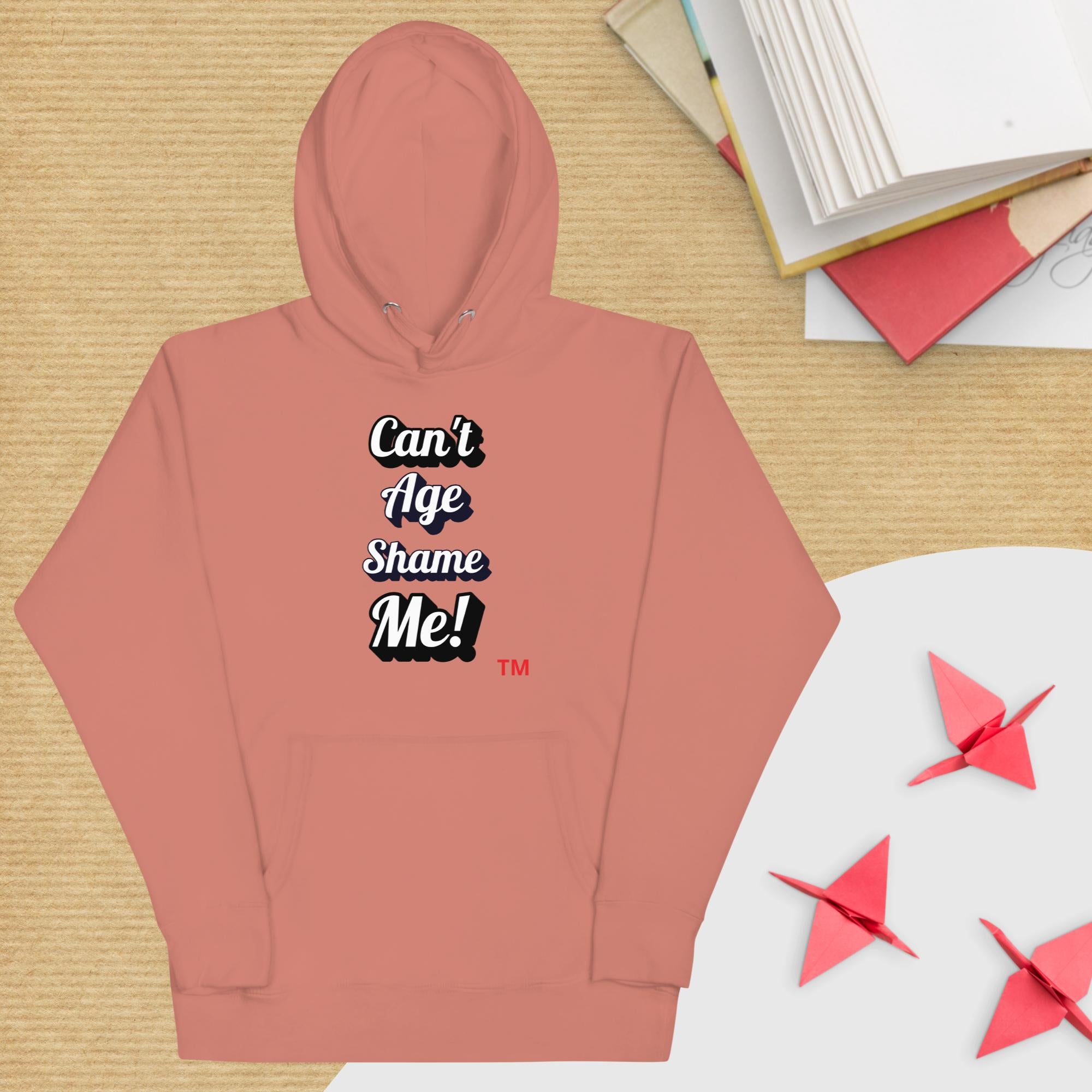 Pink hoodie with text 'Can't Age Shame Me!' on a wooden surface with books and paper cranes.