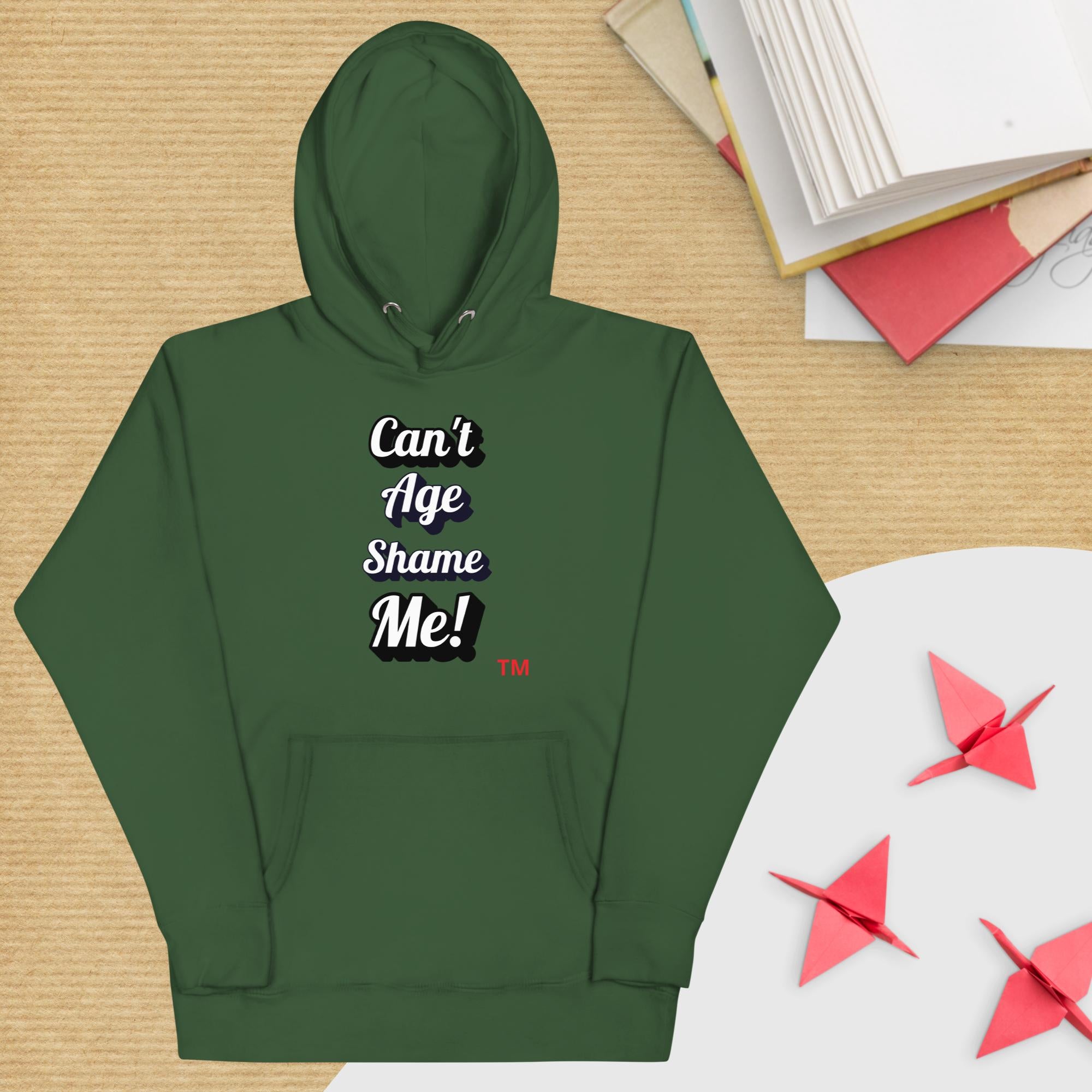 Green hoodie with text 'Can't Age Shame Me!' on a wooden surface with books and paper cranes.