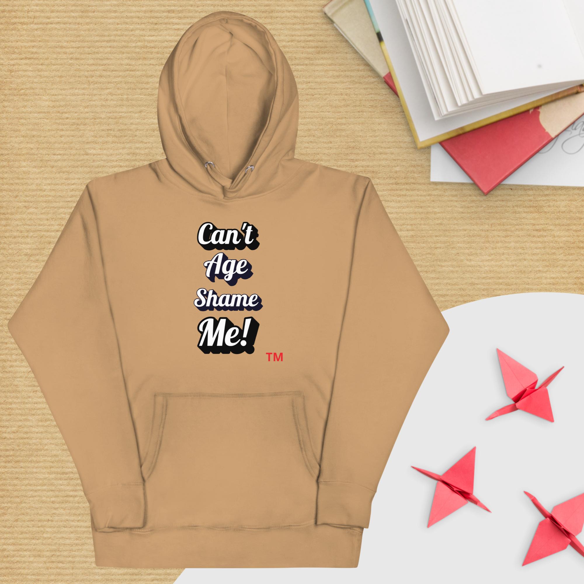 Brown hoodie with text 'Can't Age Shame Me!' on a wooden surface with books and paper cranes.