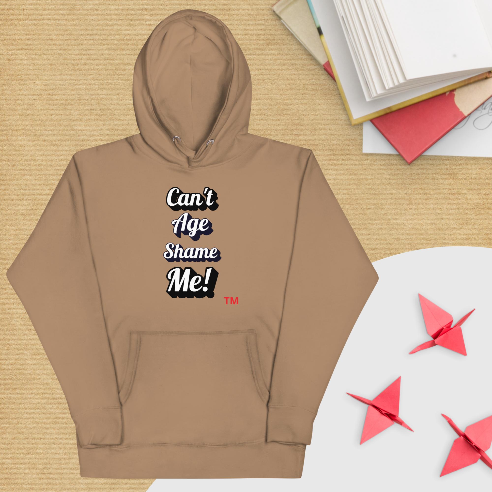 Brown hoodie with text 'Can't Age Shame Me!' on a wooden surface with books and paper cranes.
