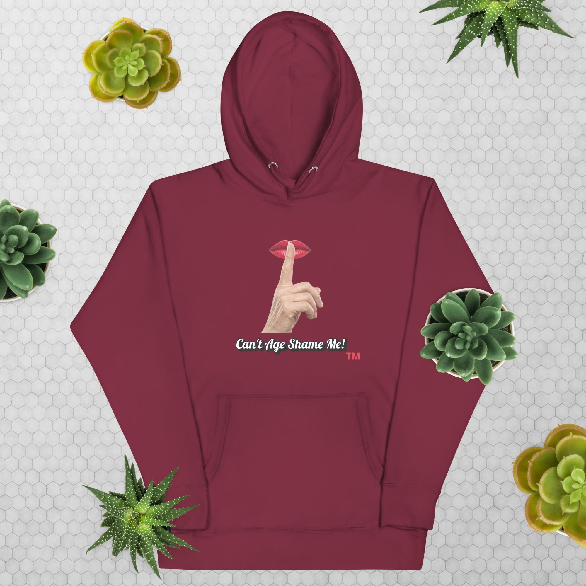 Maroon hoodie with graphic design and text, Can’t Age Shame Me!, on a patterned background with succulents