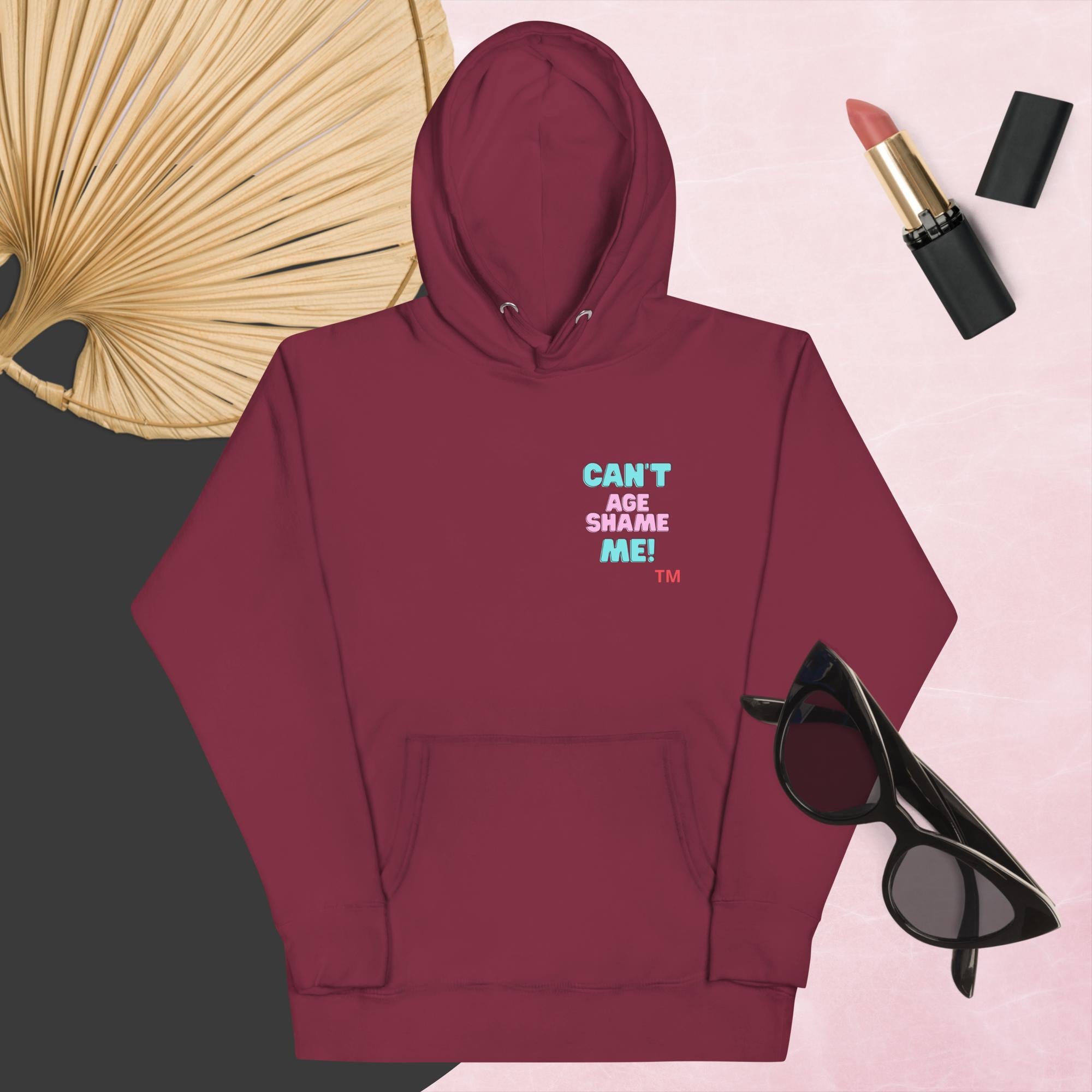 Maroon hoodie with text, 'Can't Age Shame Me' with sunglasses, lipstick, and a decorative leaf on a pink background