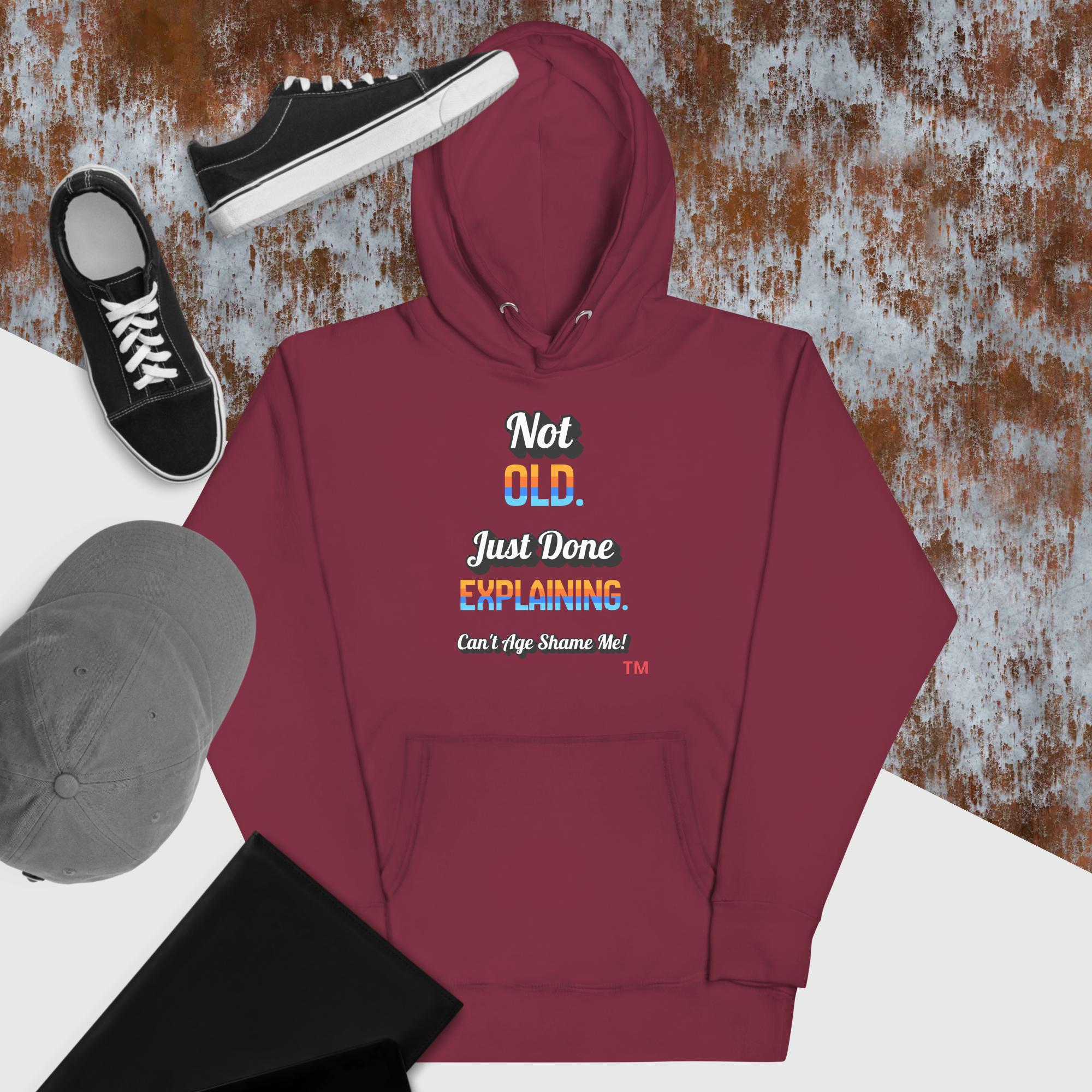 Maroon hoodie with humorous text design, black sneakers, and gray cap on a textured surface.