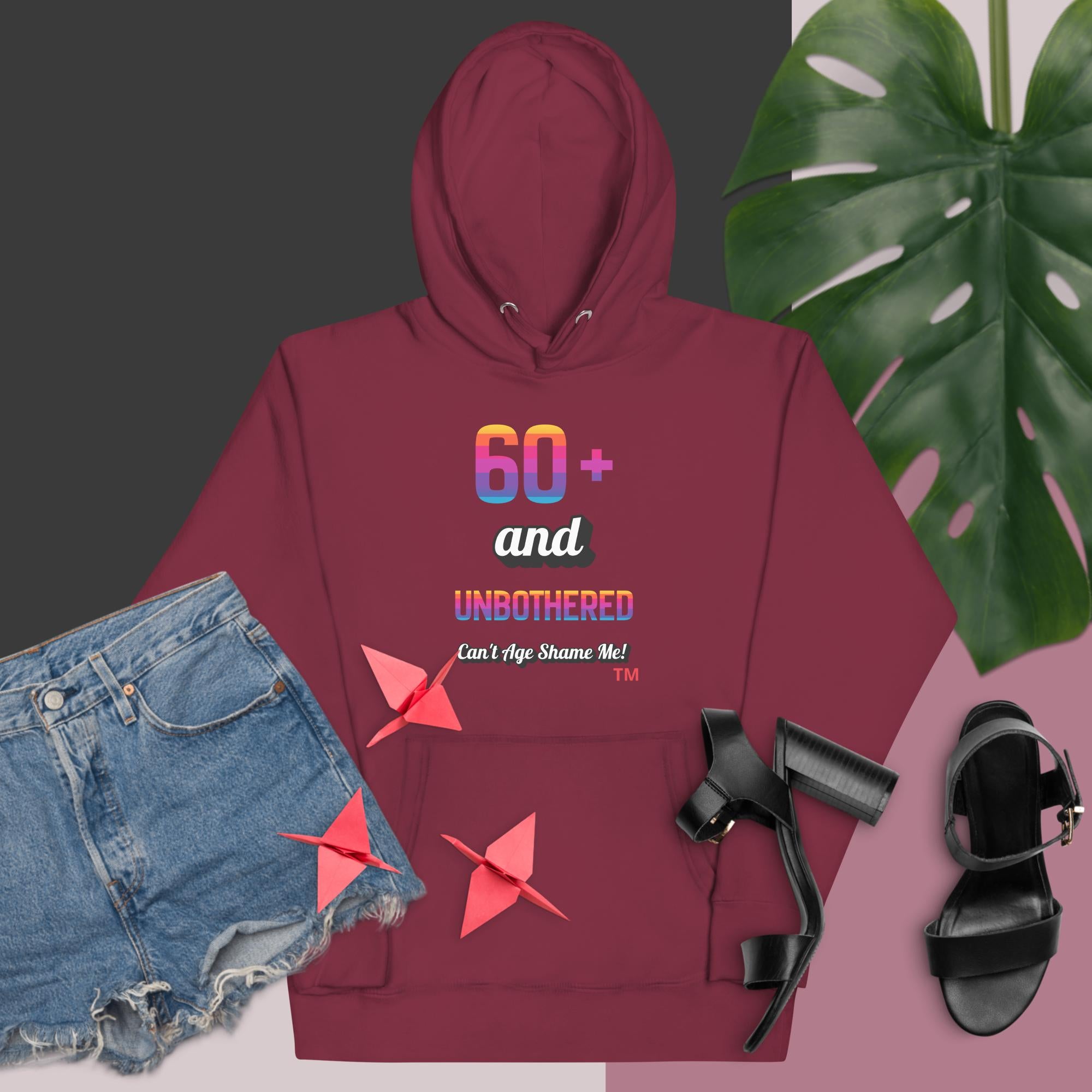 Maroon hoodie with colorful text, denim shorts, black sandals, and a monstera leaf on a pink background