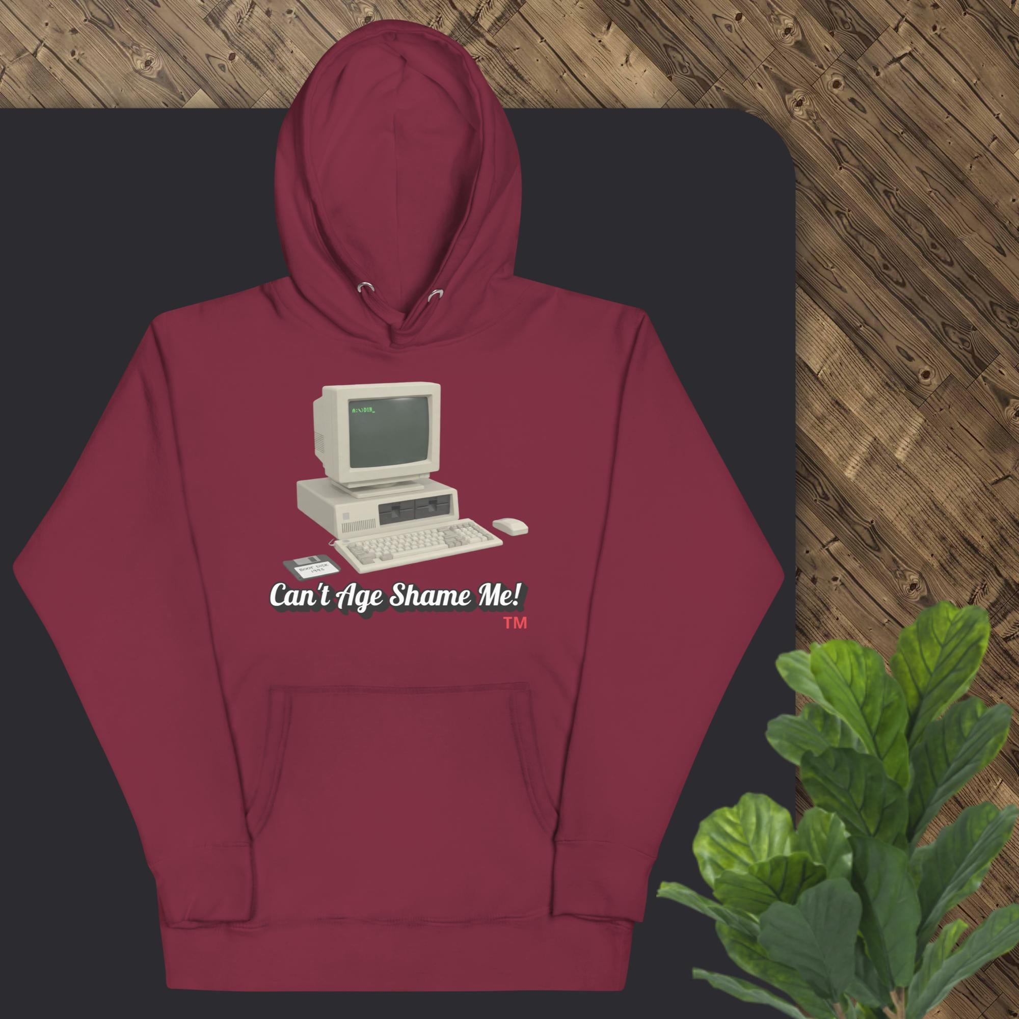 Maroon hoodie with vintage computer graphic and text on a dark surface with a plant in the corner.