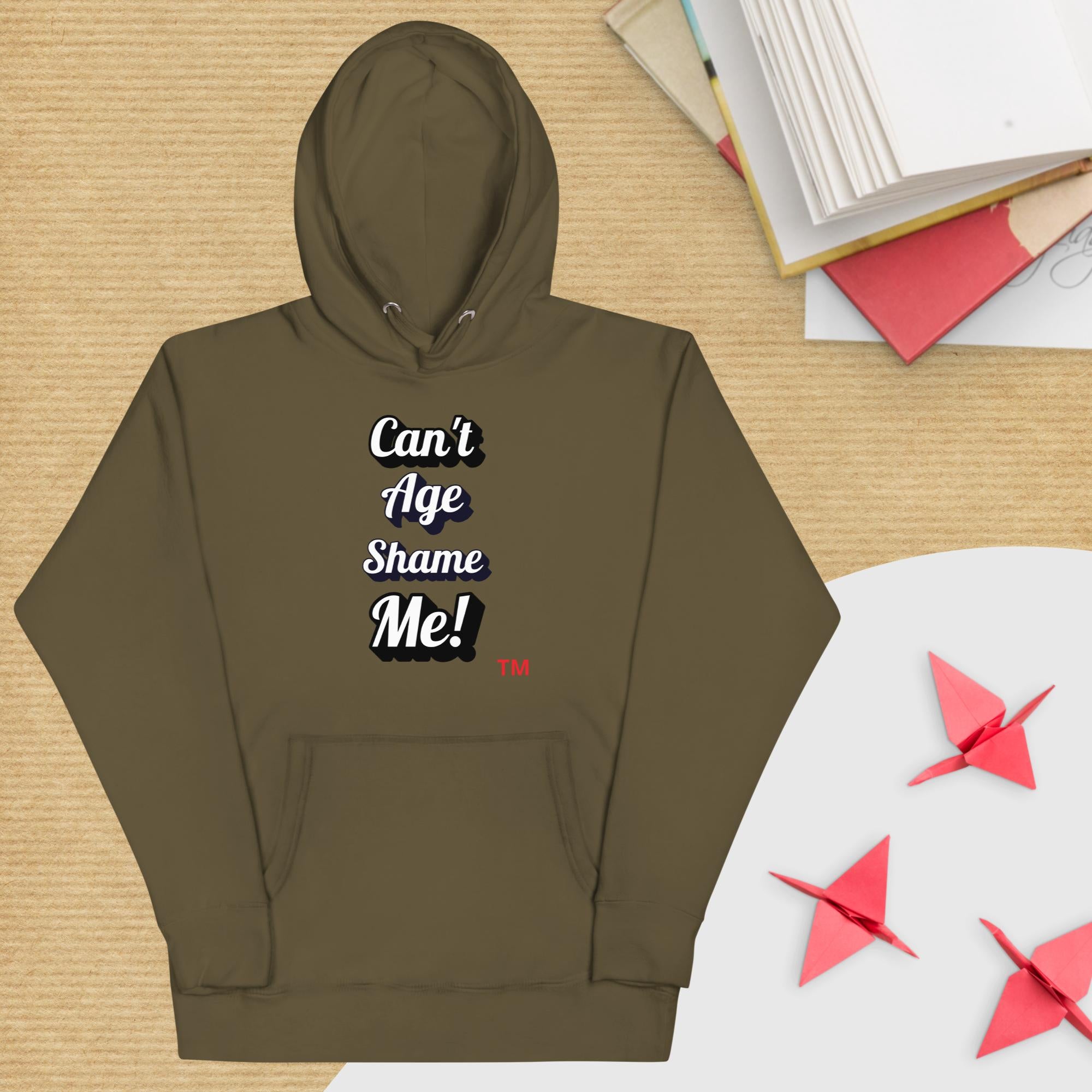 Brown hoodie with 'Can't Age Shame Me!' text on a wooden surface with books and paper cranes.