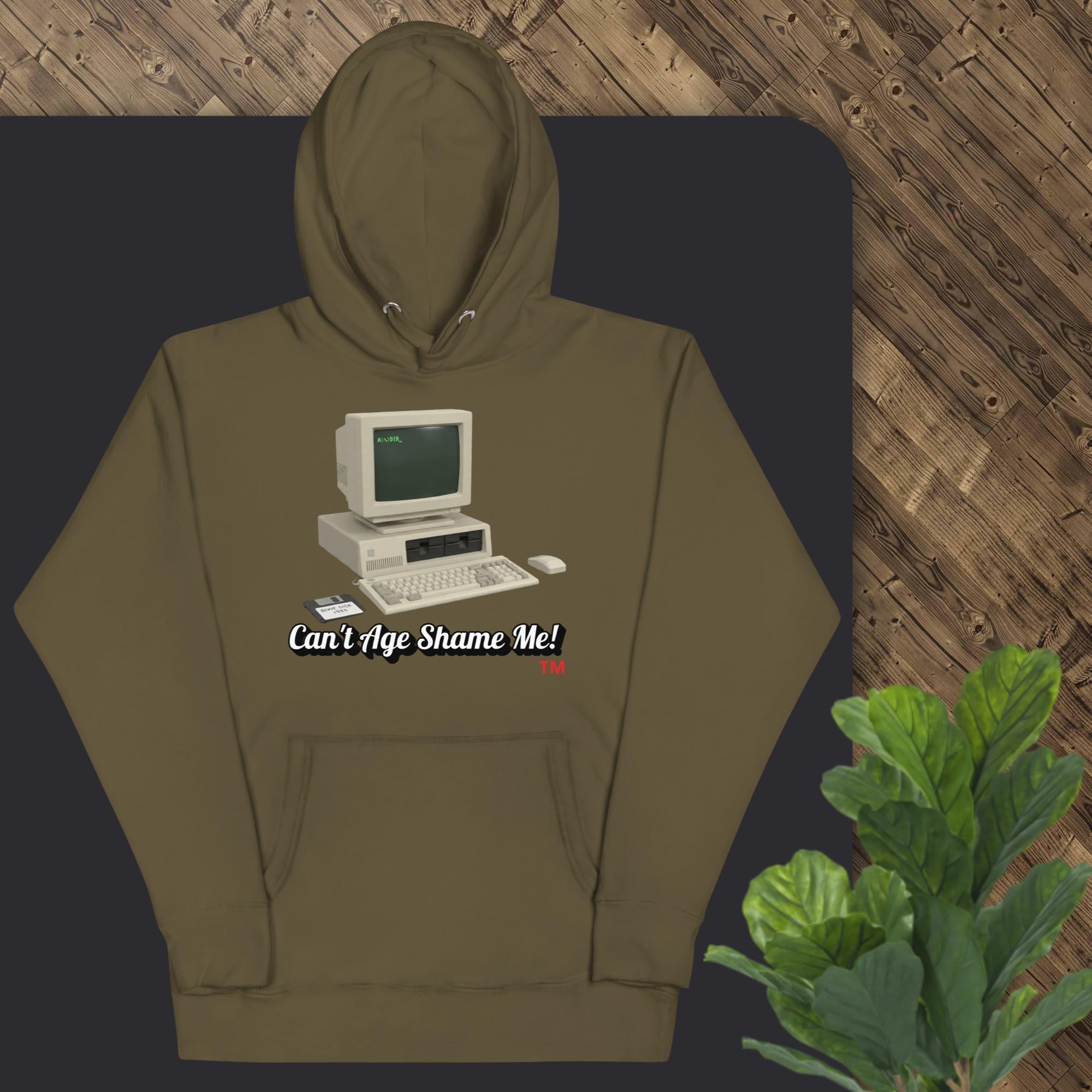 military green hoodie with vintage computer graphic and text 'Can't Age Shame Me' on a dark surface.