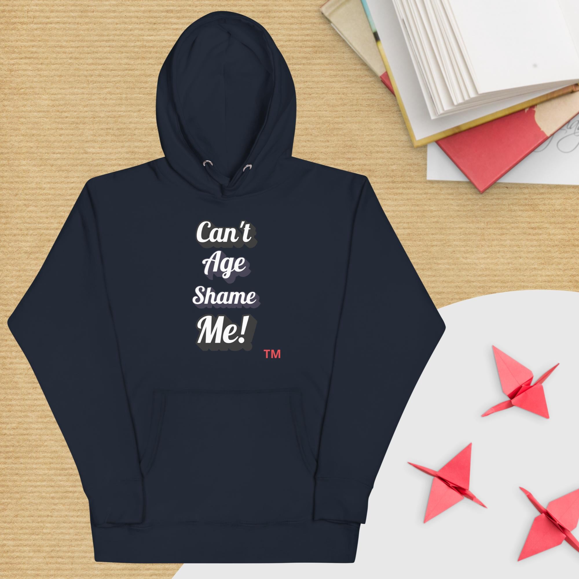 Navy blue hoodie with 'Can't Age Shame Me!' text on a wooden surface with books and paper cranes.