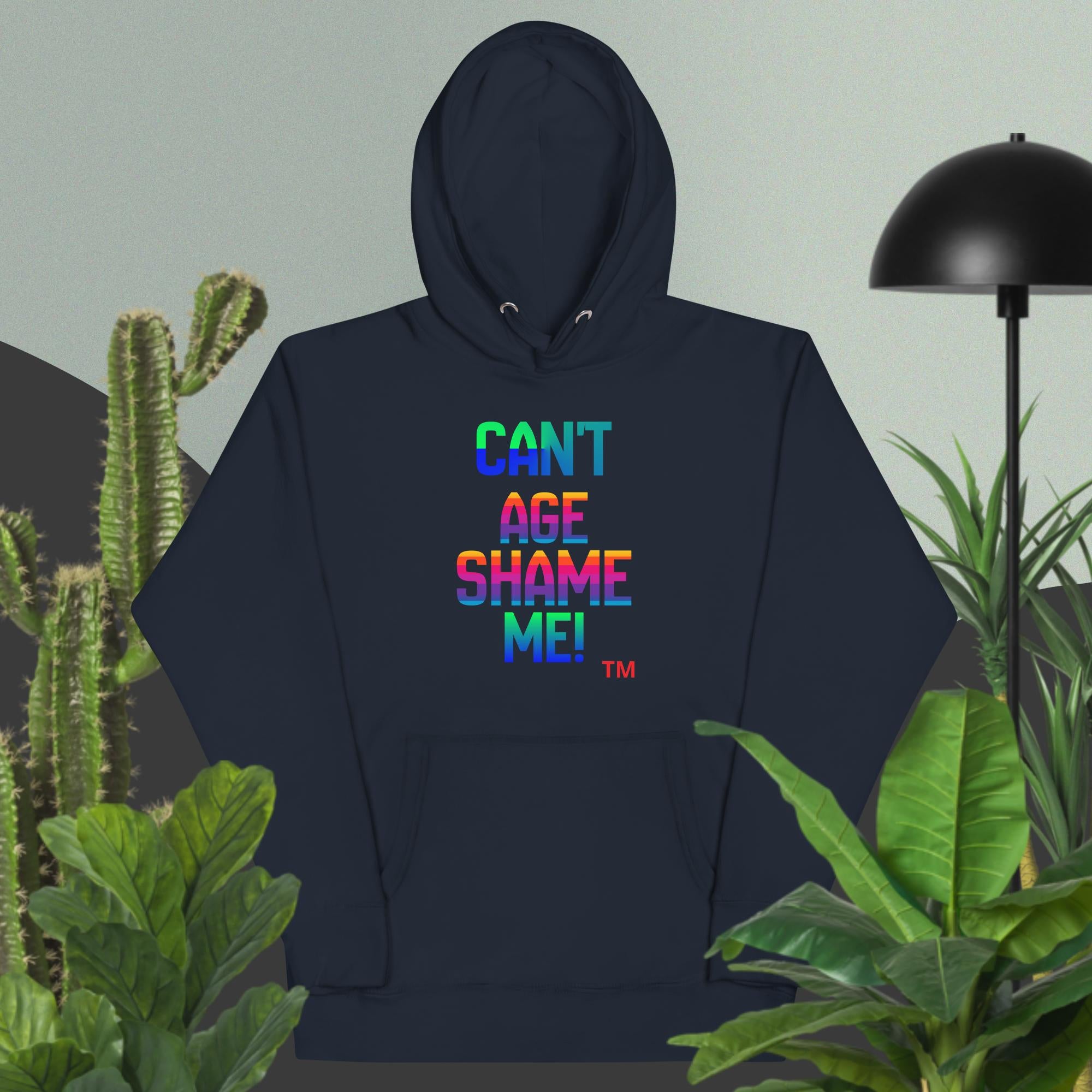 Navy blue hoodie with colorful text 'CAN'T AGE SHAME ME!' in a room with plants and a lamp.