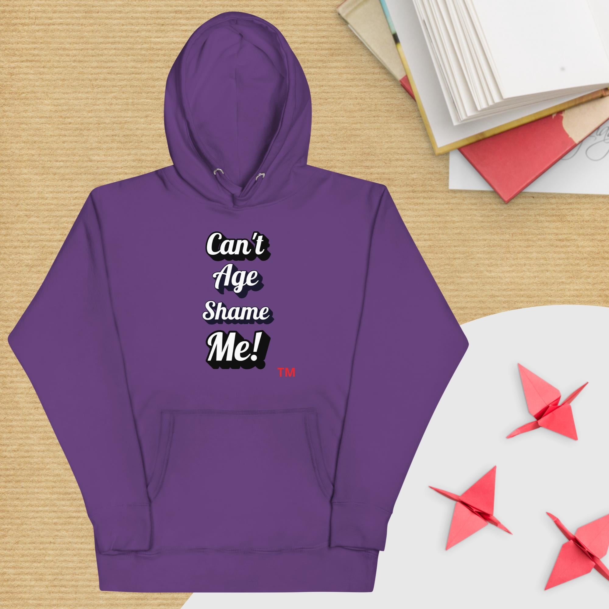 Purple hoodie with text 'Can't Age Shame Me!' on a wooden surface with books and paper cranes.