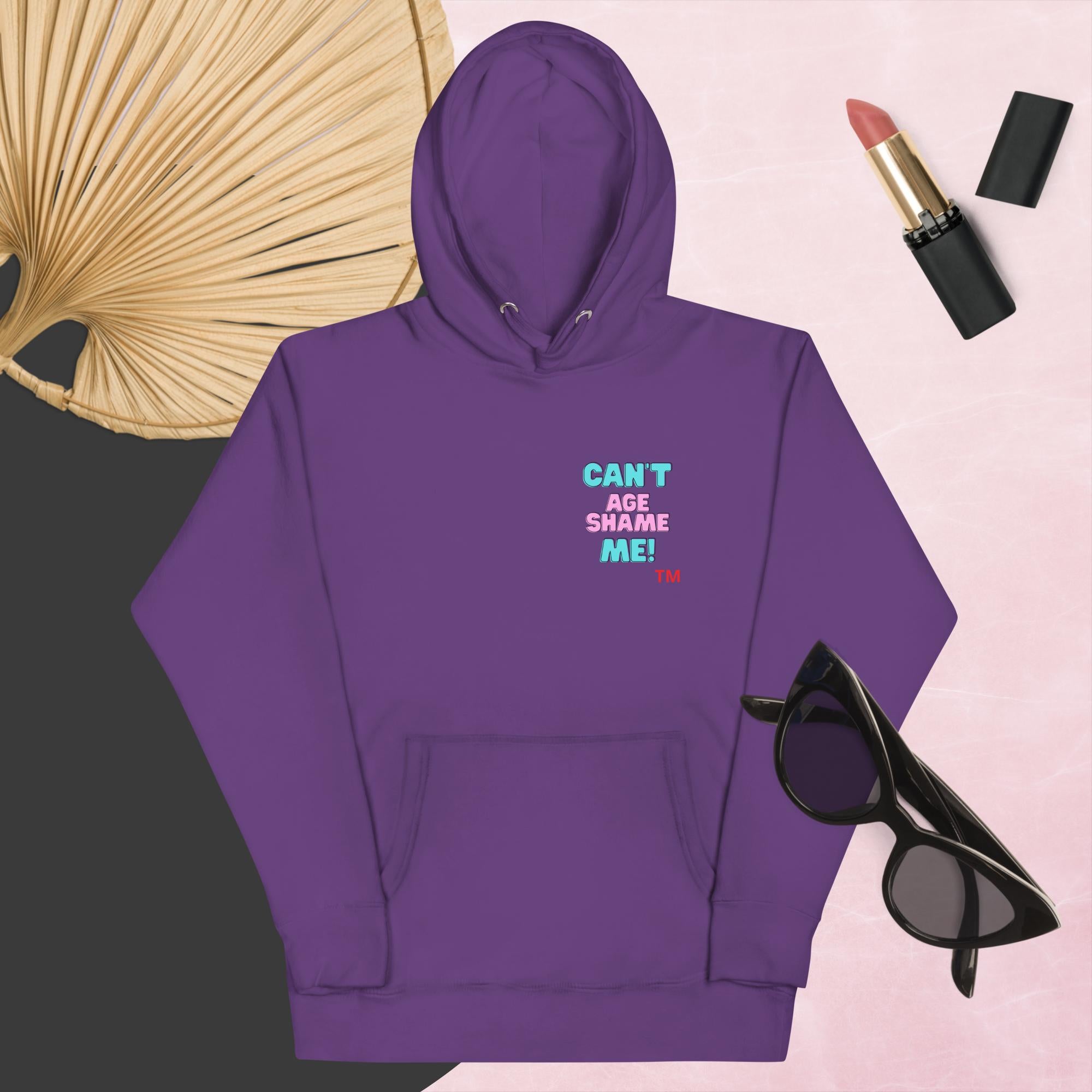 Purple hoodie with text, 'Can't Age Shame Me' with sunglasses, lipstick, and a decorative leaf on a pink background