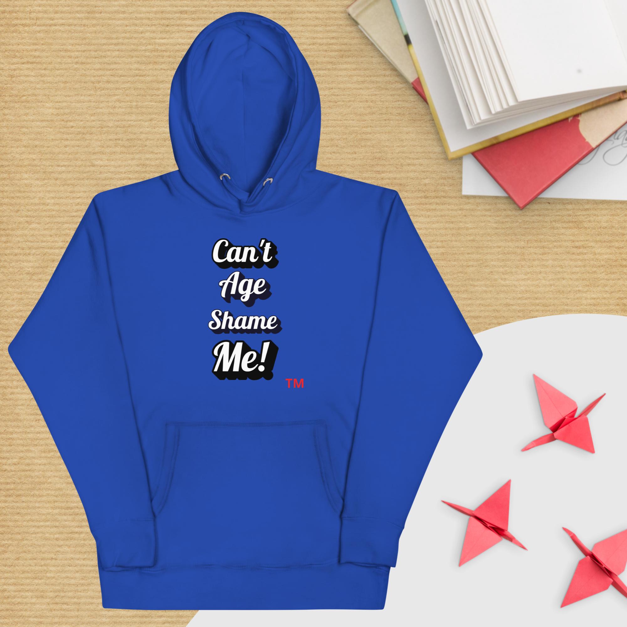 Blue hoodie with 'Can't Age Shame Me!' text on a wooden surface with books and paper cranes.