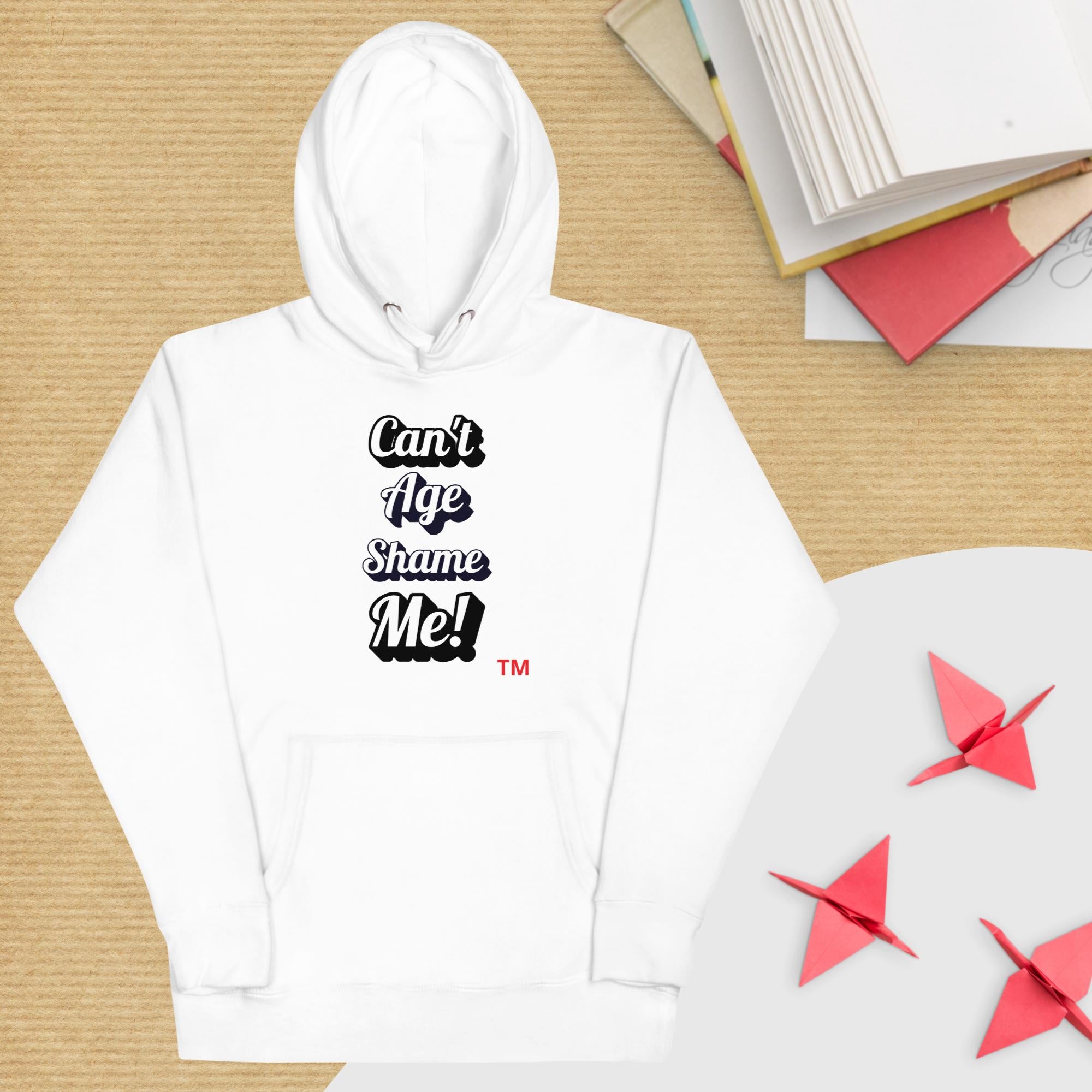 White hoodie with text 'Can't Age Shame Me!' on a wooden surface with books and paper stars.