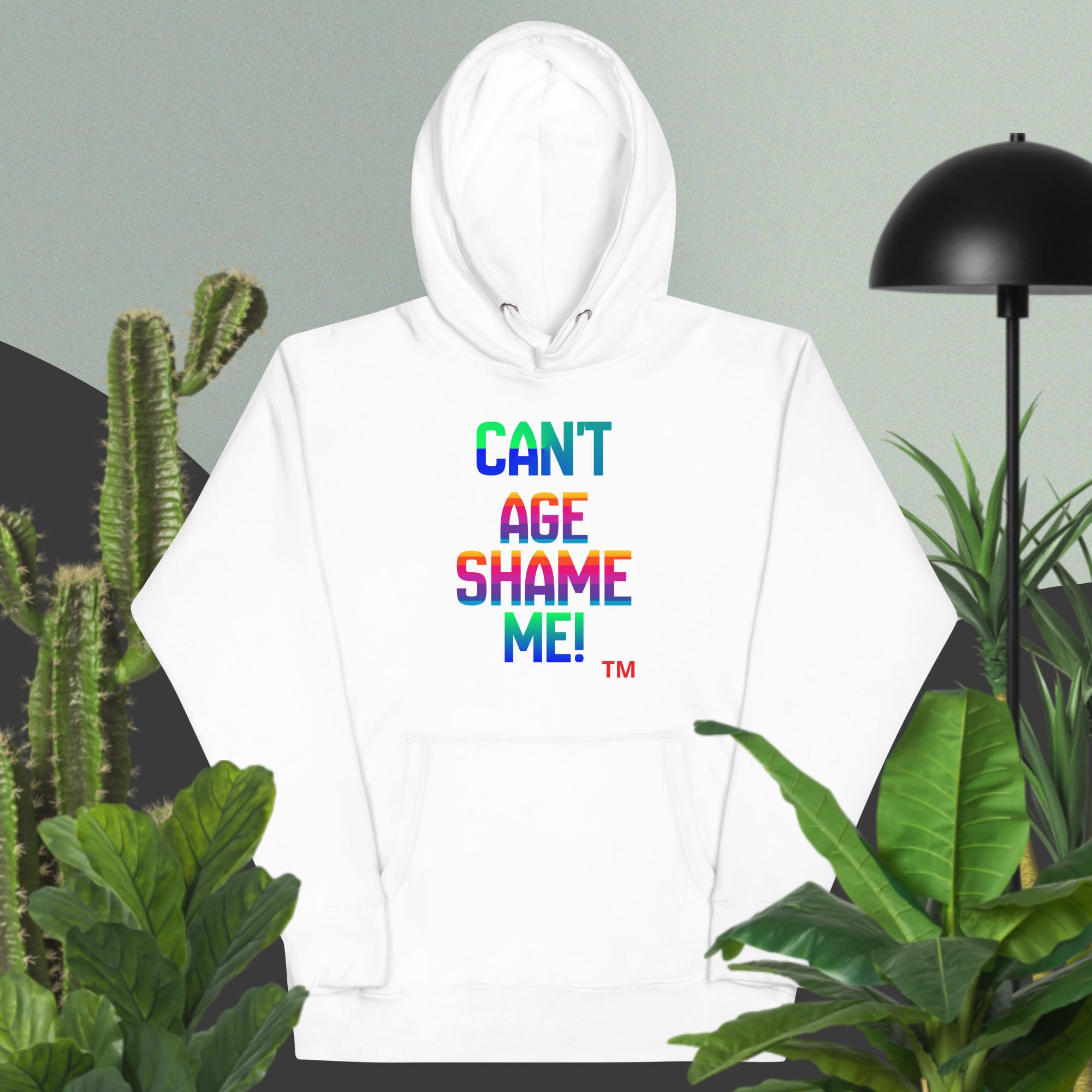 White hoodie with colorful text 'CANT AGE SHAME ME!' surrounded by plants and a lamp.