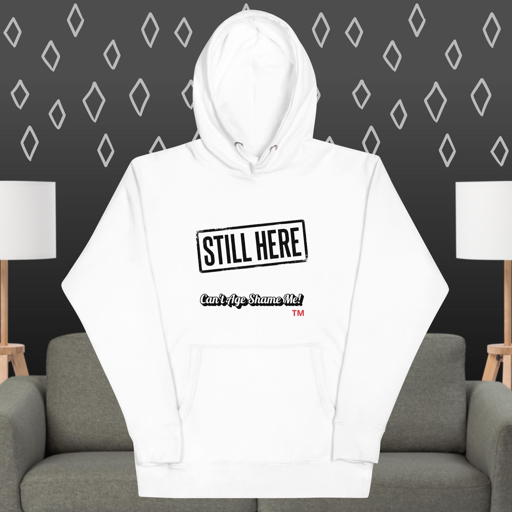 White hoodie with 'STILL HERE' text on a gray couch against a patterned wall.