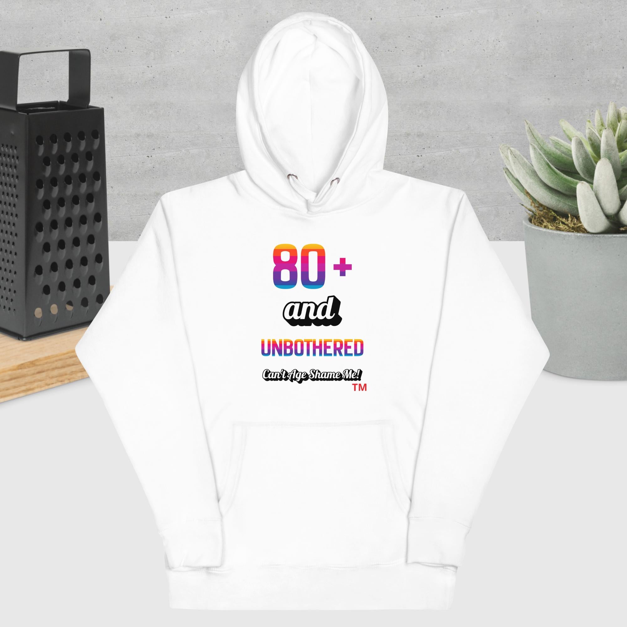 White hoodie with colorful text design on a gray surface with a black grater and plant in the background