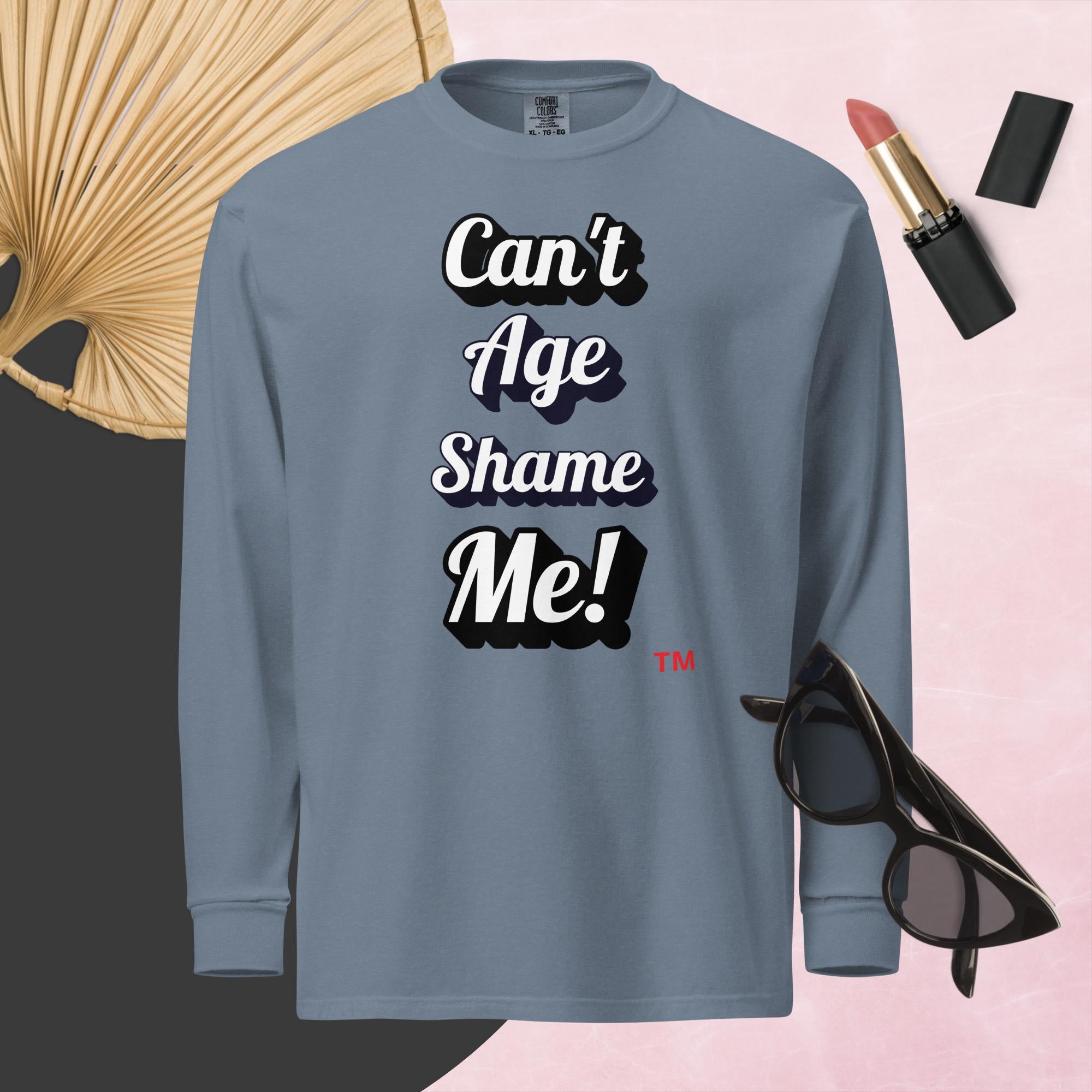 Gray long-sleeve shirt with 'Can't Age Shame Me!' text, surrounded by sunglasses, lipstick, and a fan on a pink background.