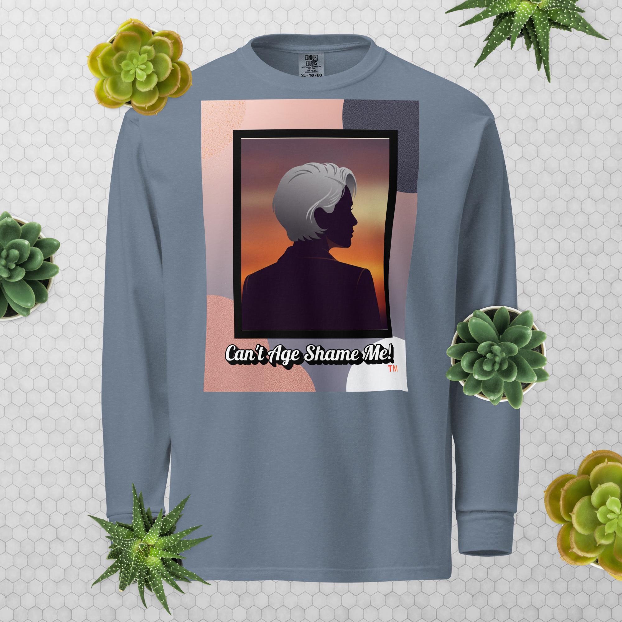 Blue Jean long-sleeve shirt with a graphic design featuring a woman with gray hair silhouette and text: Can’t Age Shame Me! surrounded by decorative plants on a light gray background.

