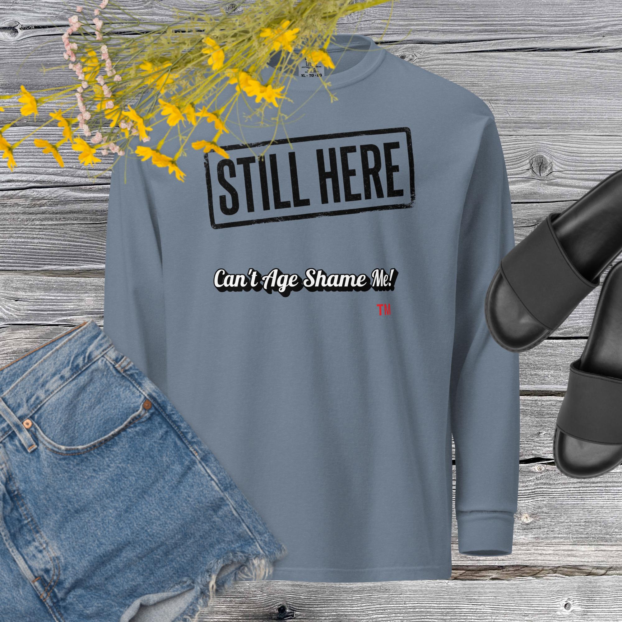 Blue  Jean sweatshirt with 'STILL HERE Can't Age Shame Me' text on a wooden surface with jeans and sandals.