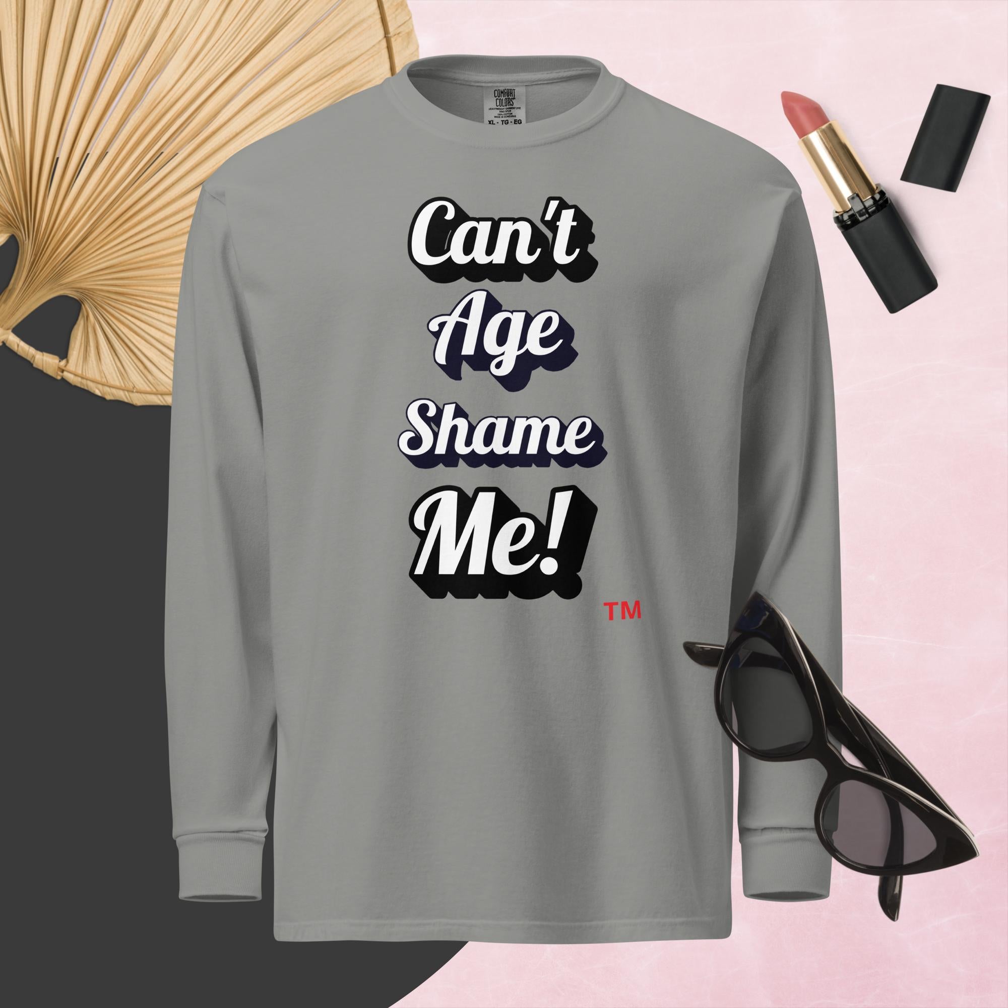 Gray long-sleeve shirt with 'Can't Age Shame Me!' text, surrounded by sunglasses, lipstick, and a decorative leaf on a pink background.
