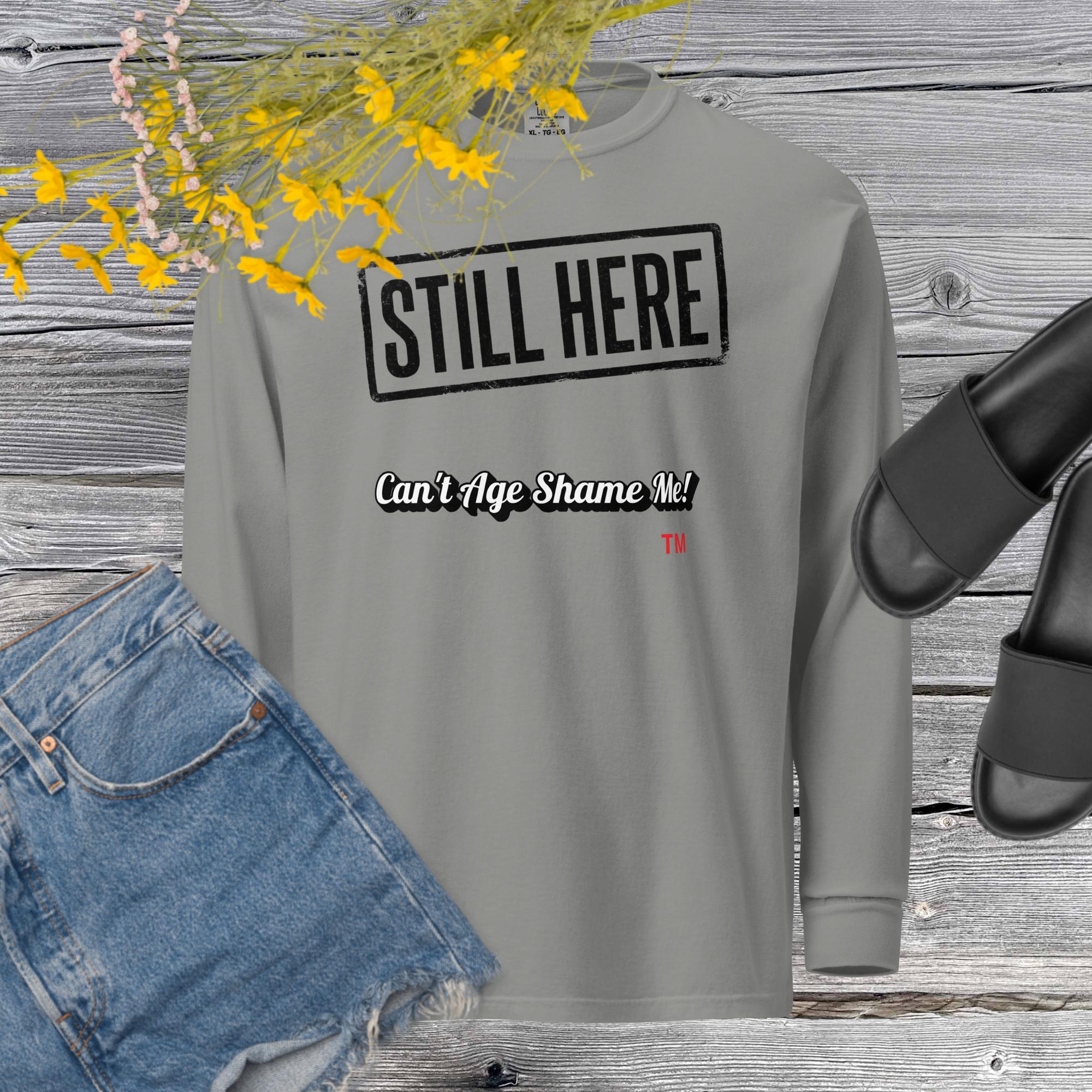 Gray sweatshirt with text 'STILL HERE Can't Age Shame Me' on a wooden surface with denim jeans and sandals.