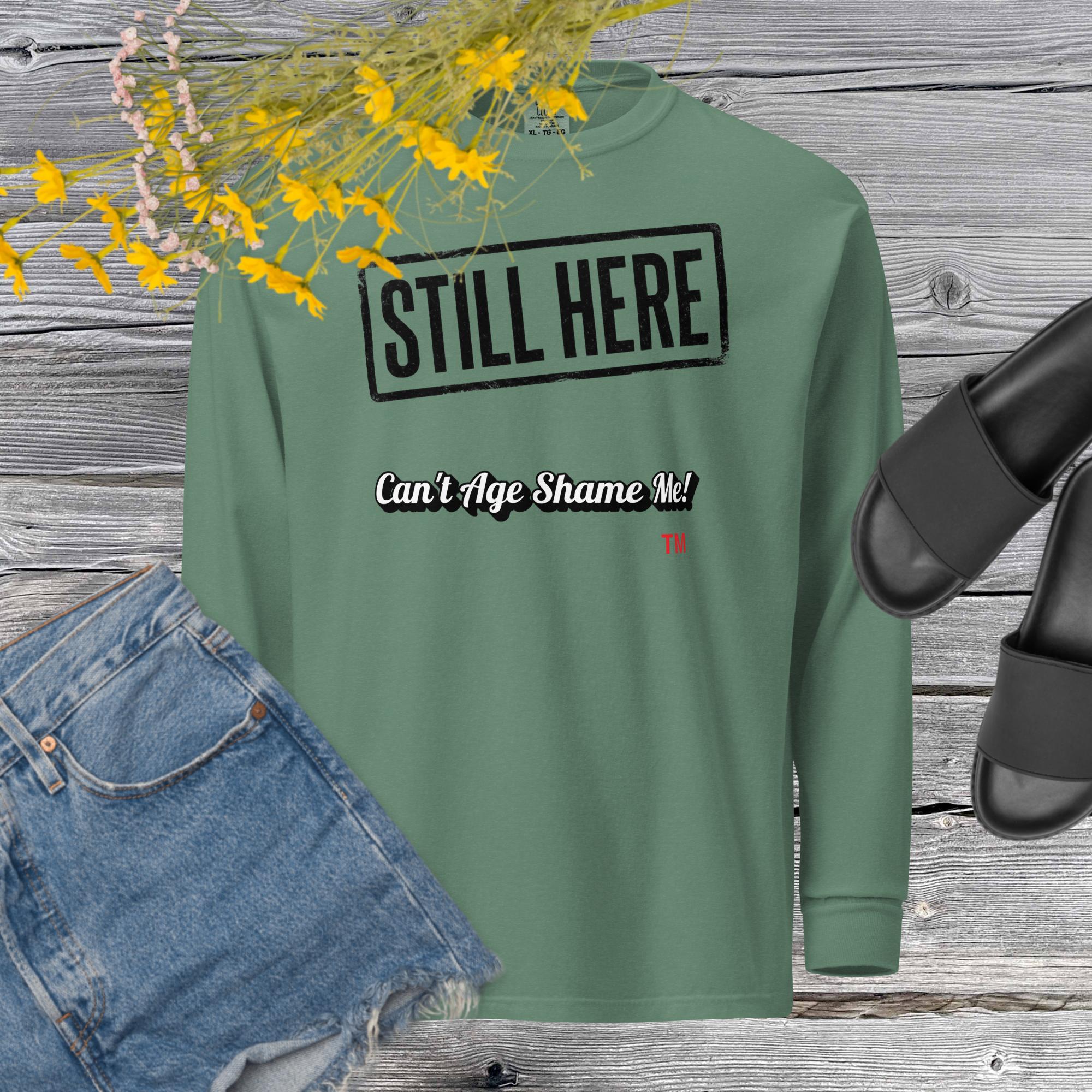 Green sweatshirt with 'STILL HERE Can't Age Shame Me' text on a wooden surface with jeans and sandals.