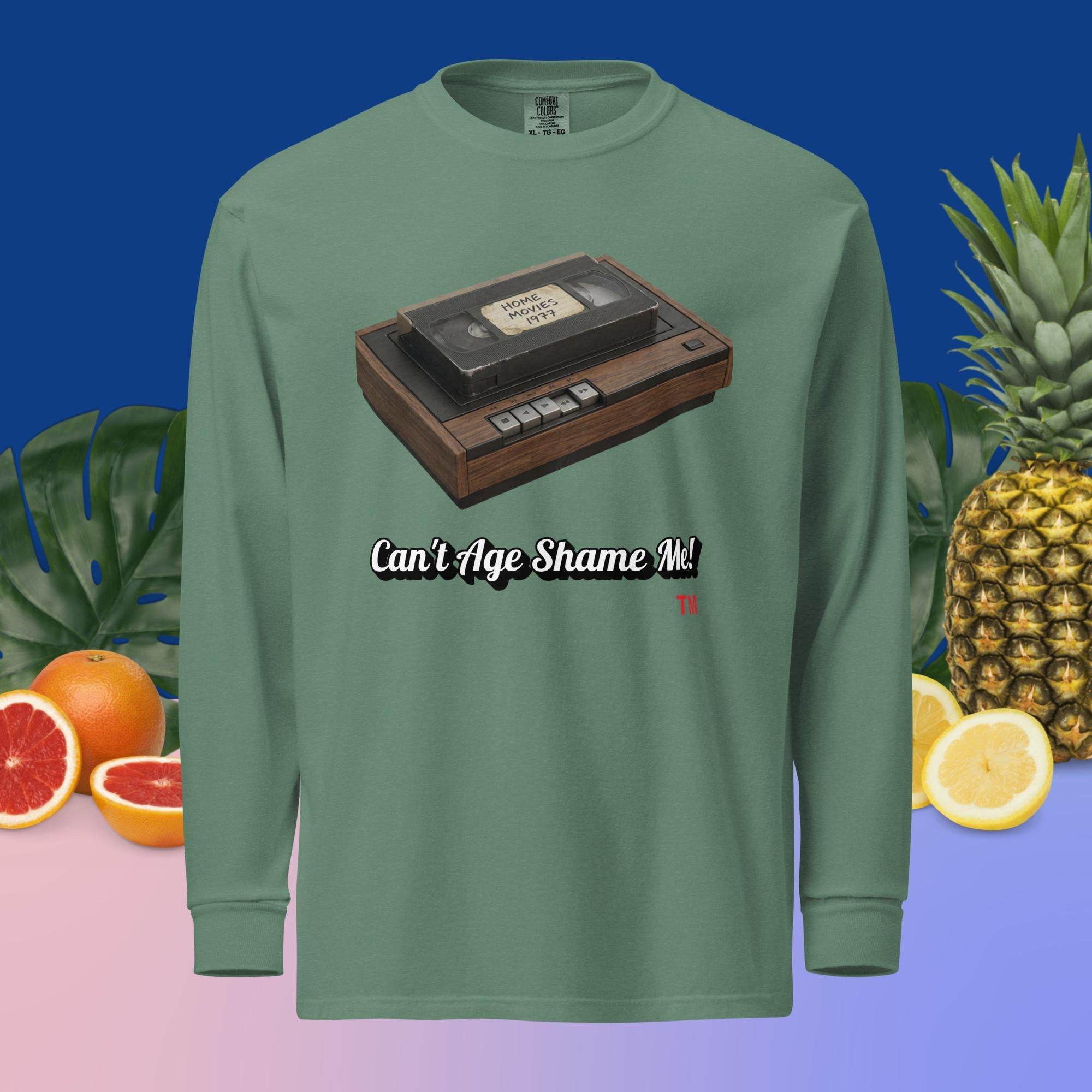 Green long-sleeve shirt with a cassette tape graphic and text 'Can't Age Shame Me!' on a colorful background with fruits and plants.