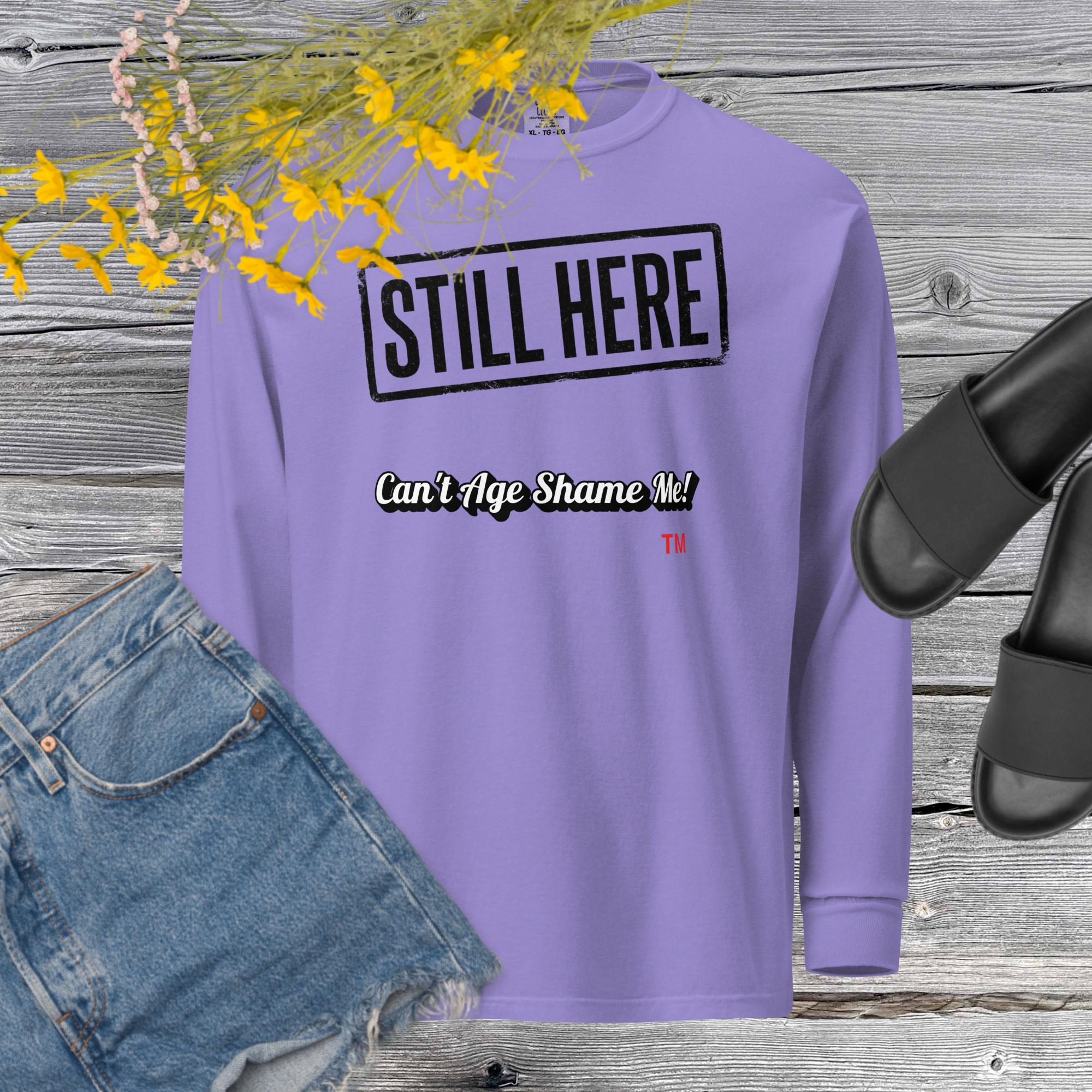Violet sweatshirt with 'STILL HERE Can't Age Shame Me' text on a wooden surface with jeans and sandals.
