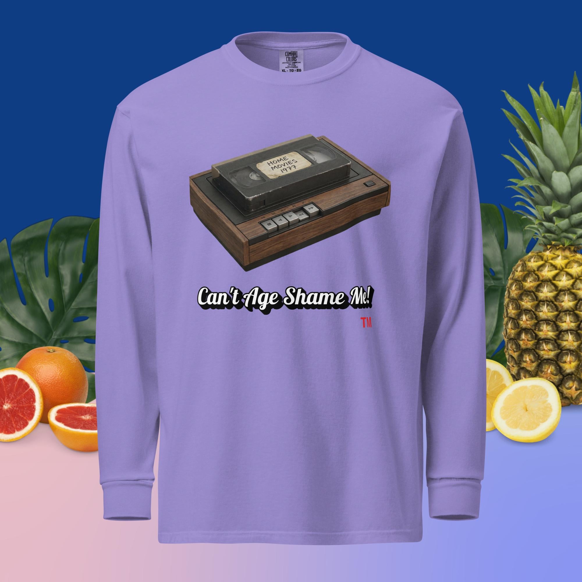 violet long-sleeve shirt with a VHS tape design and text can't age shame me, surrounded by fruits and plants.
