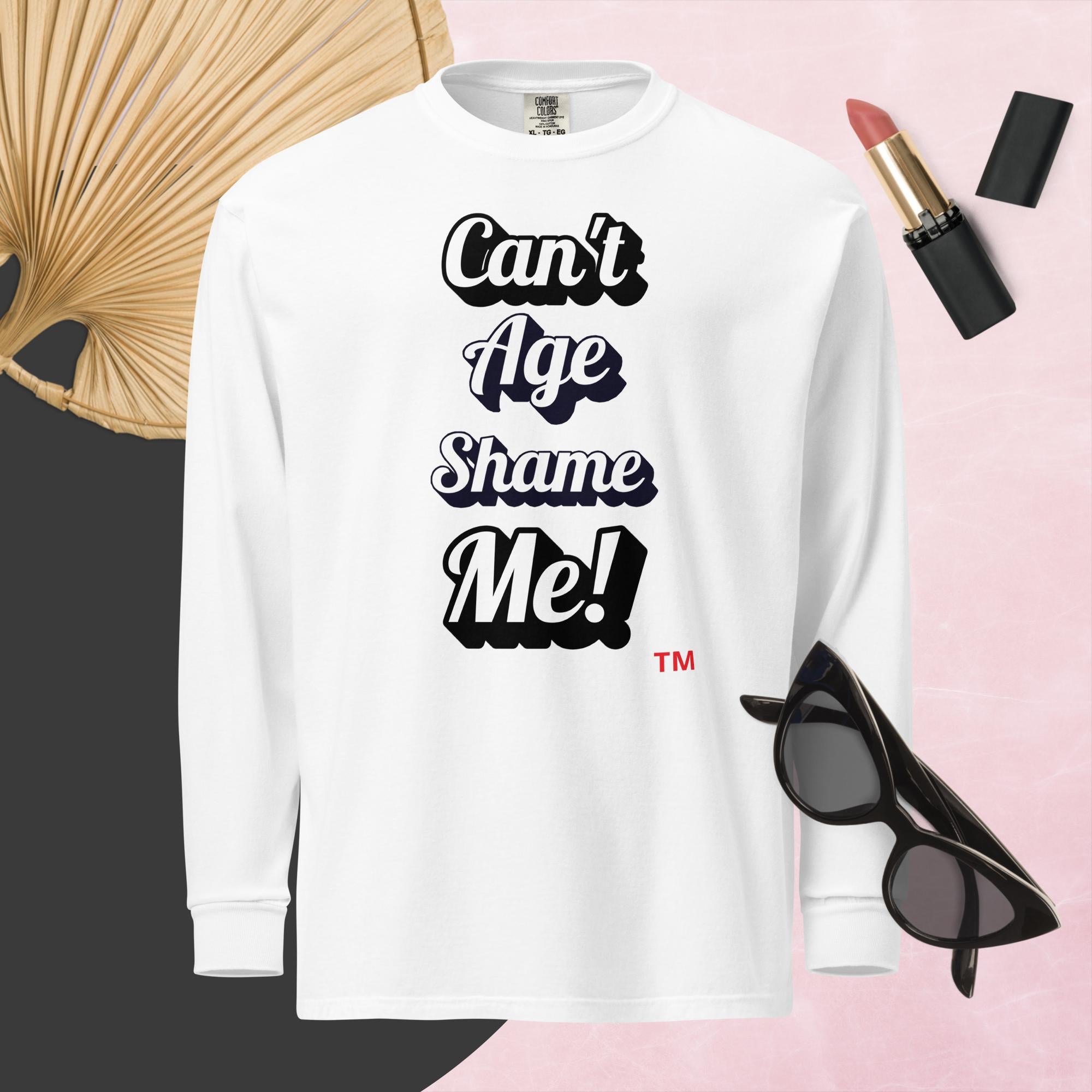 White long-sleeve shirt with 'Can't Age Shame Me!' text, sunglasses, lipstick, and decorative item on a pink background