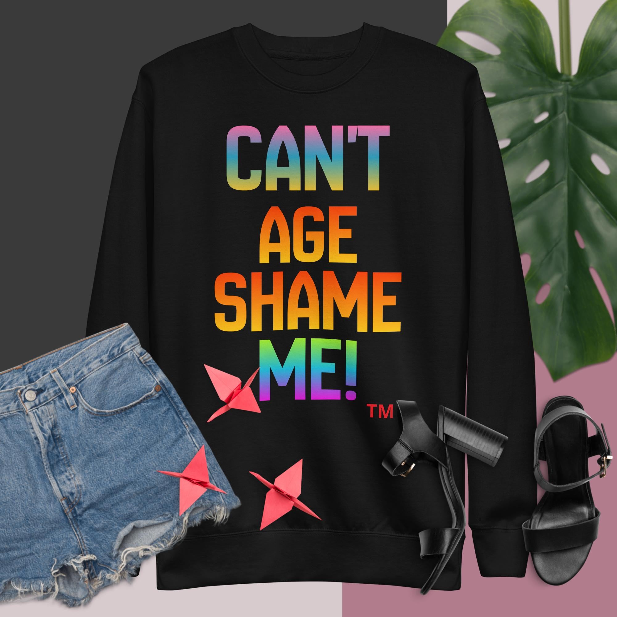 Black sweatshirt with colorful text 'CAN'T AGE SHAME ME!' displayed with denim shorts and black sandals.