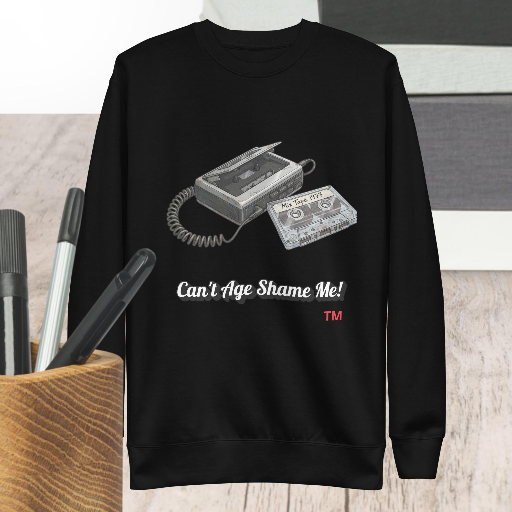 Black sweatshirt with cassette tape design and text 'Can't Age Shame Me!' on a wooden surface.