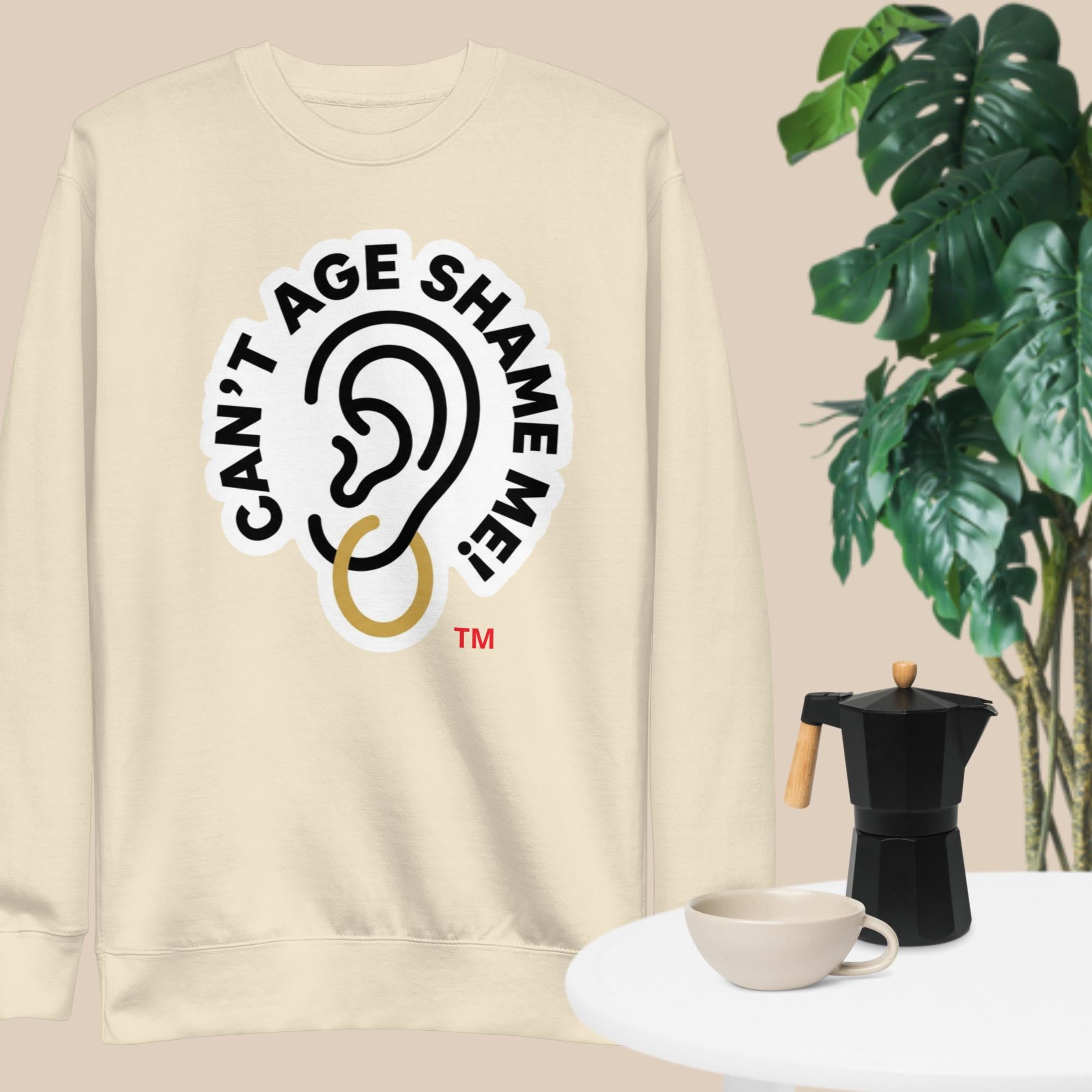 Bone sweatshirt with 'Can't Age Shame Me' logo on a beige background with a plant and coffee set.