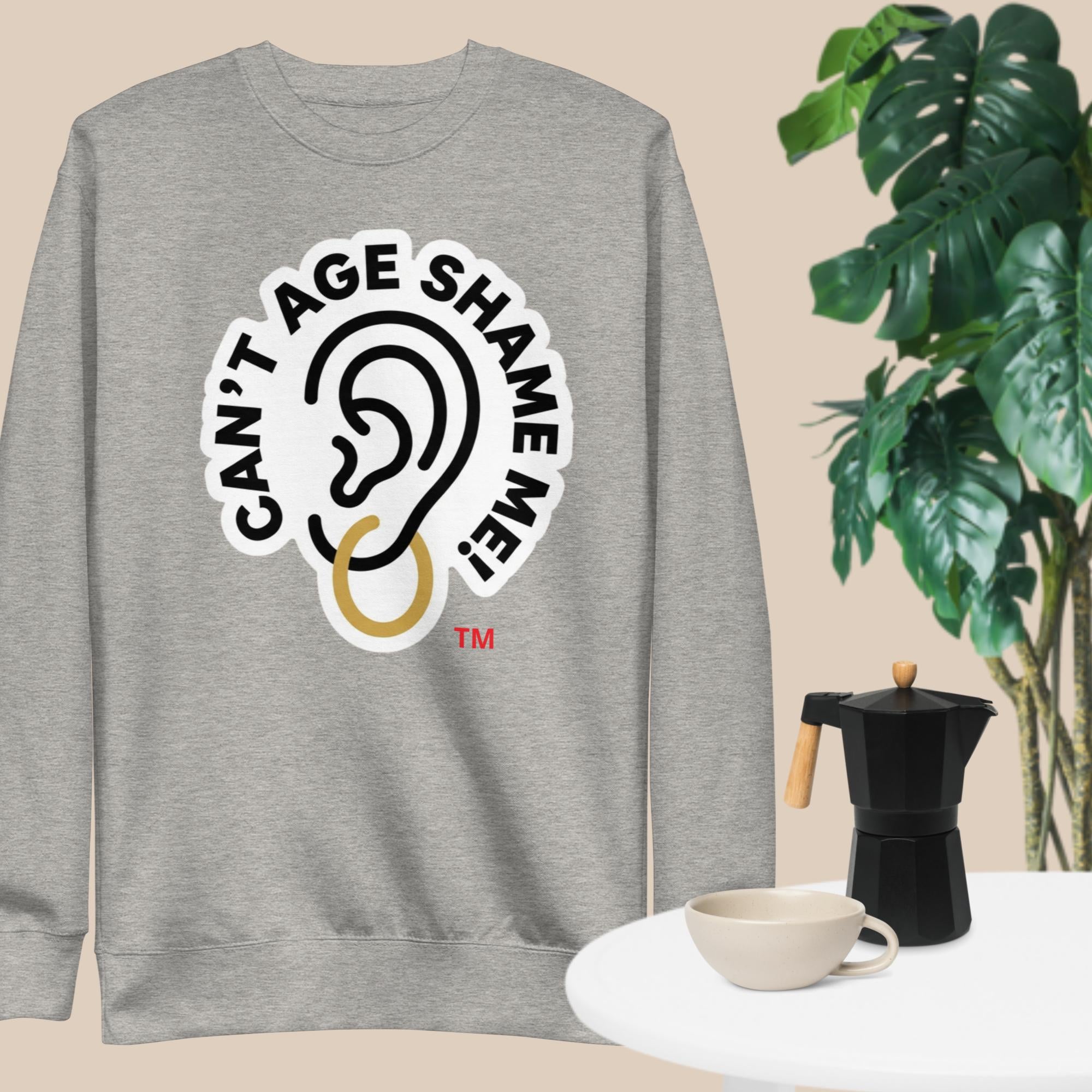 Gray sweatshirt with 'Can't Age Shame Me!' graphic on a beige background with a plant and coffee mug.