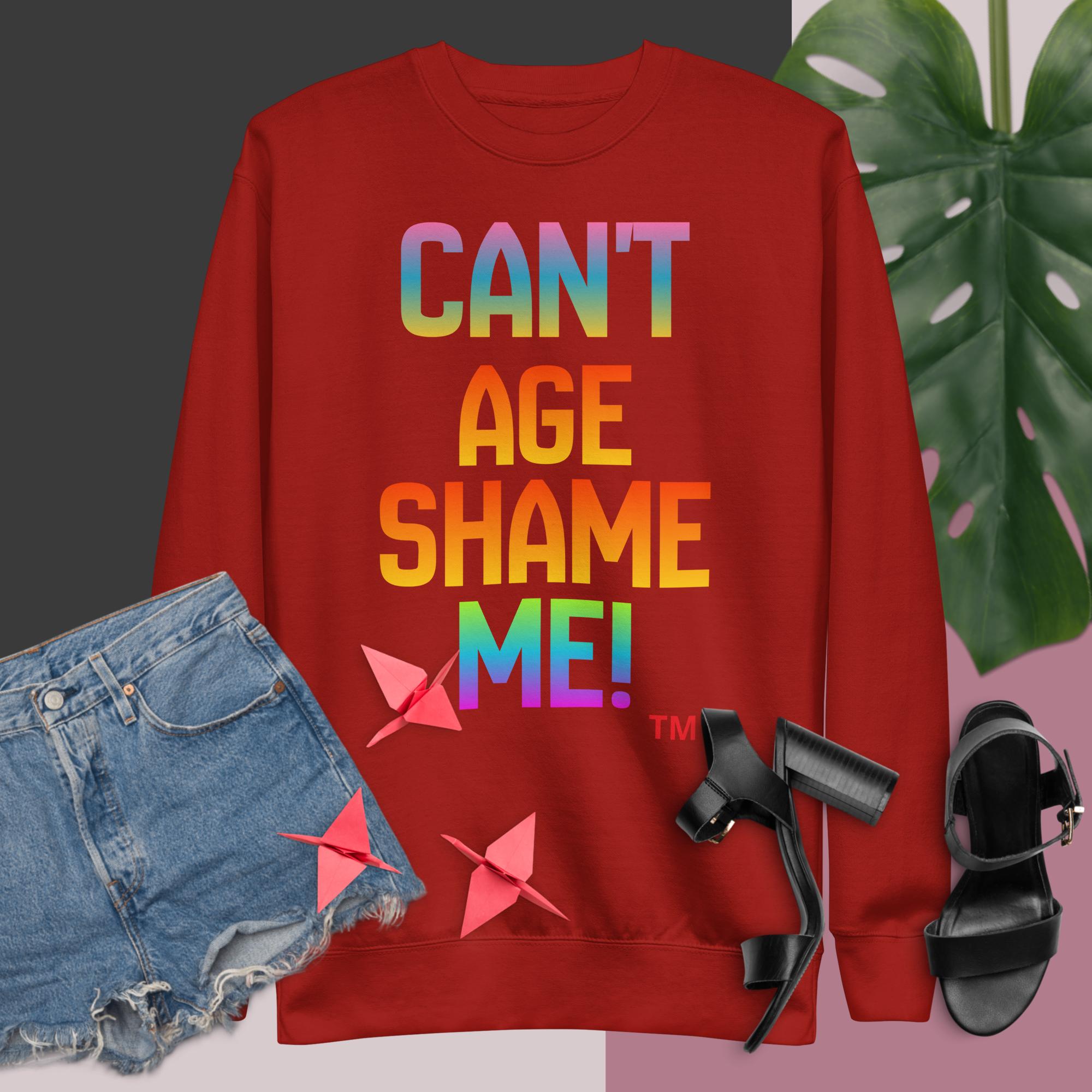 Red sweatshirt with colorful text, denim shorts, black sandals, and a plant on a pink and gray background