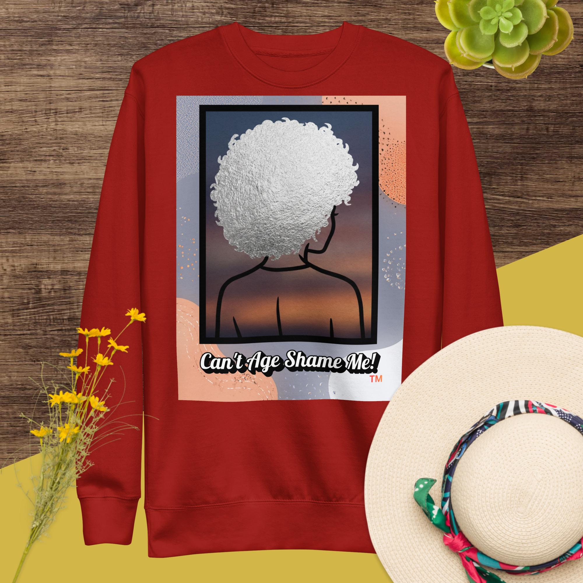 Red sweatshirt with a graphic design of a person with white textured hair and text 'Can't Age Shame Me!' on a wooden surface with flowers and a hat.