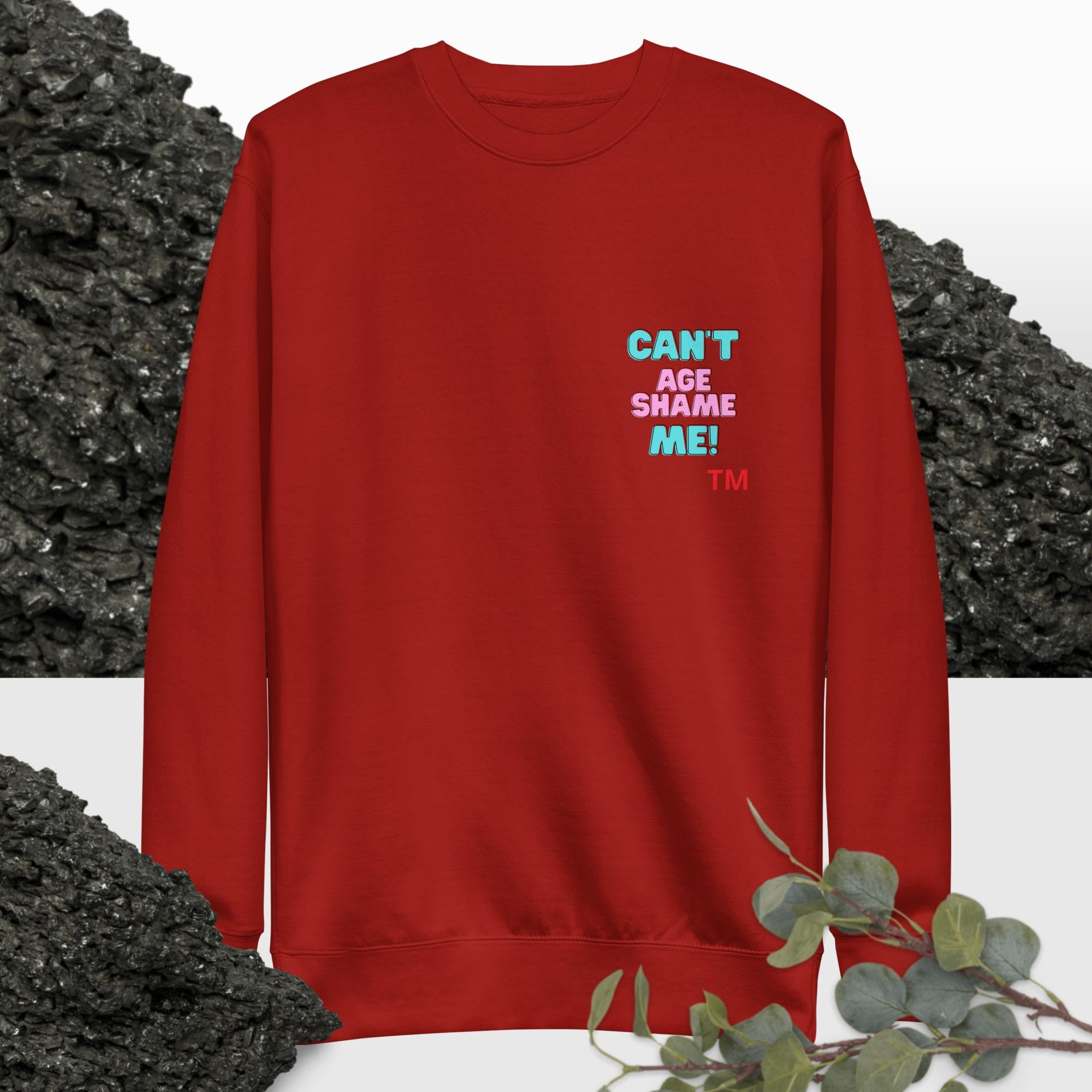 Cardinal sweatshirt with colorful text: 'Can't Age Shame Me'  on a background with black rocks and greenery.