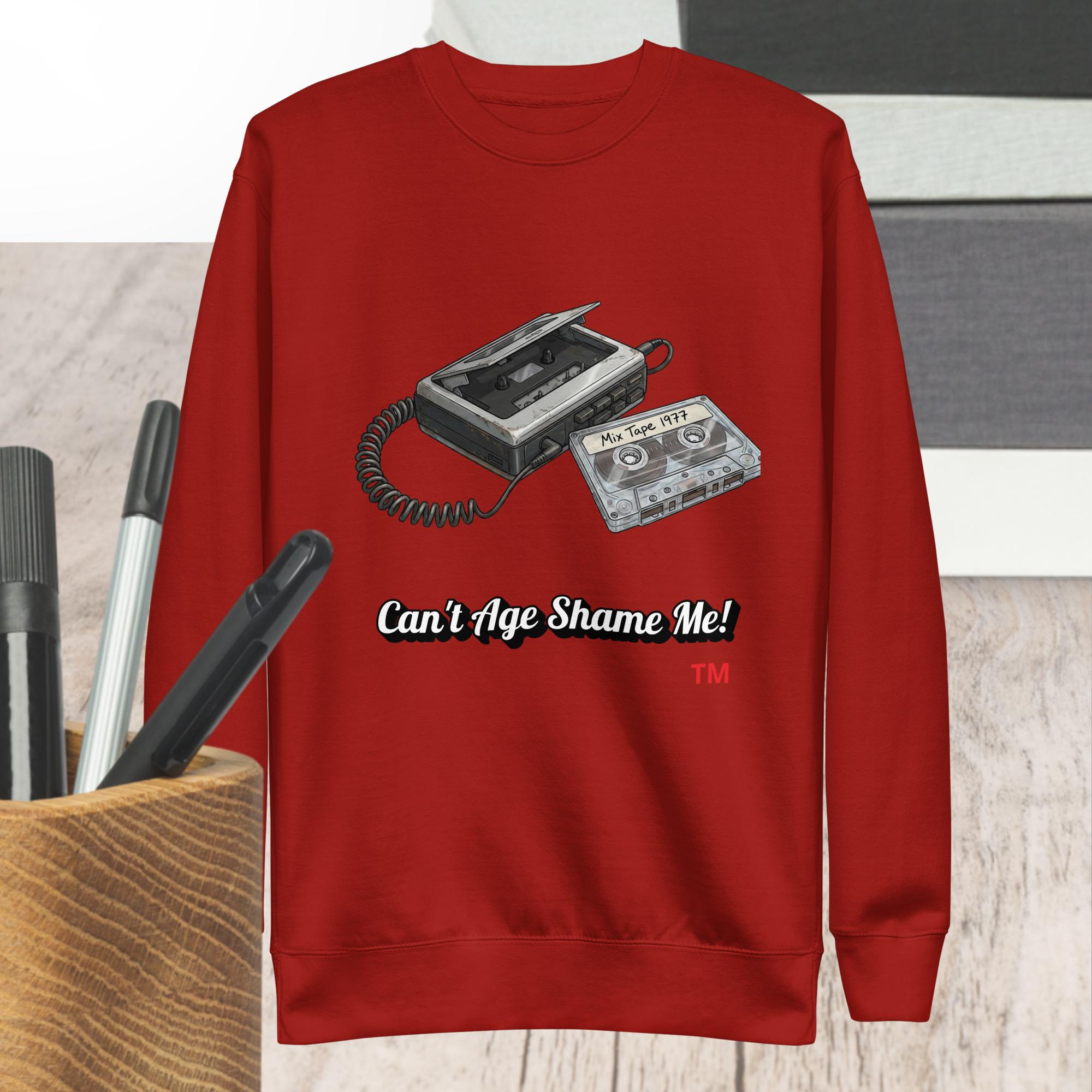 Red sweatshirt with a cassette tape design and text 'Can't Age Shame Me!' on a wooden surface.