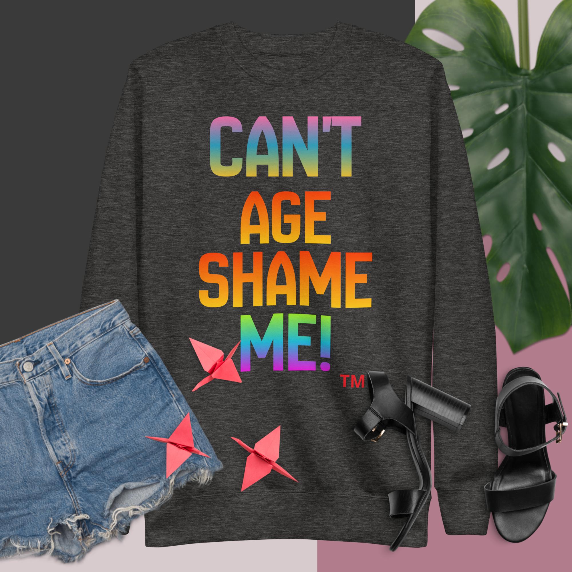 Dark gray sweatshirt with colorful text 'CAN'T AGE SHAME ME!' displayed with denim shorts, black sandals, and a plant.