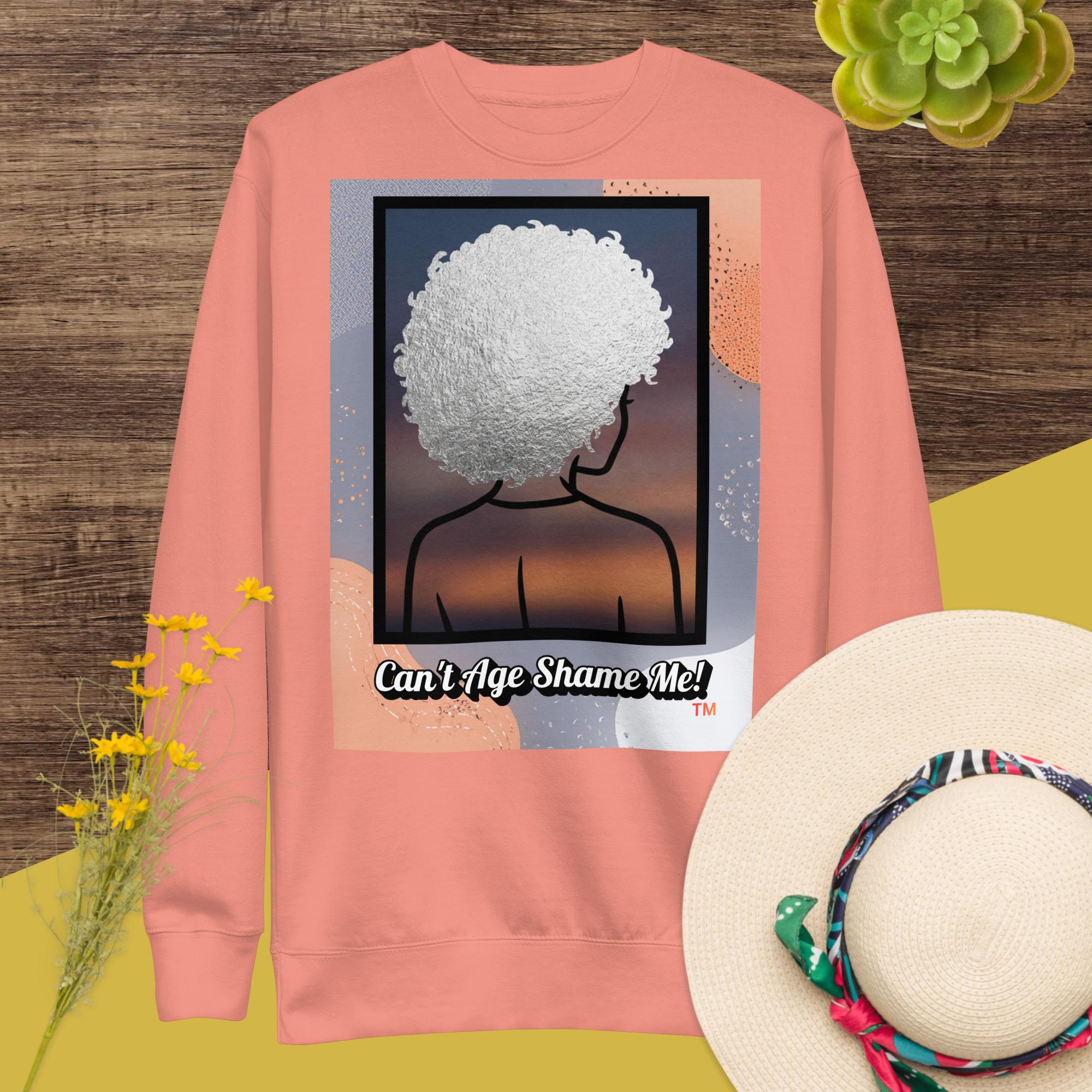 Pinkish sweatshirt with a graphic design of a person with textured hair and text 'Can't Age Shame Me!' on a colorful background.