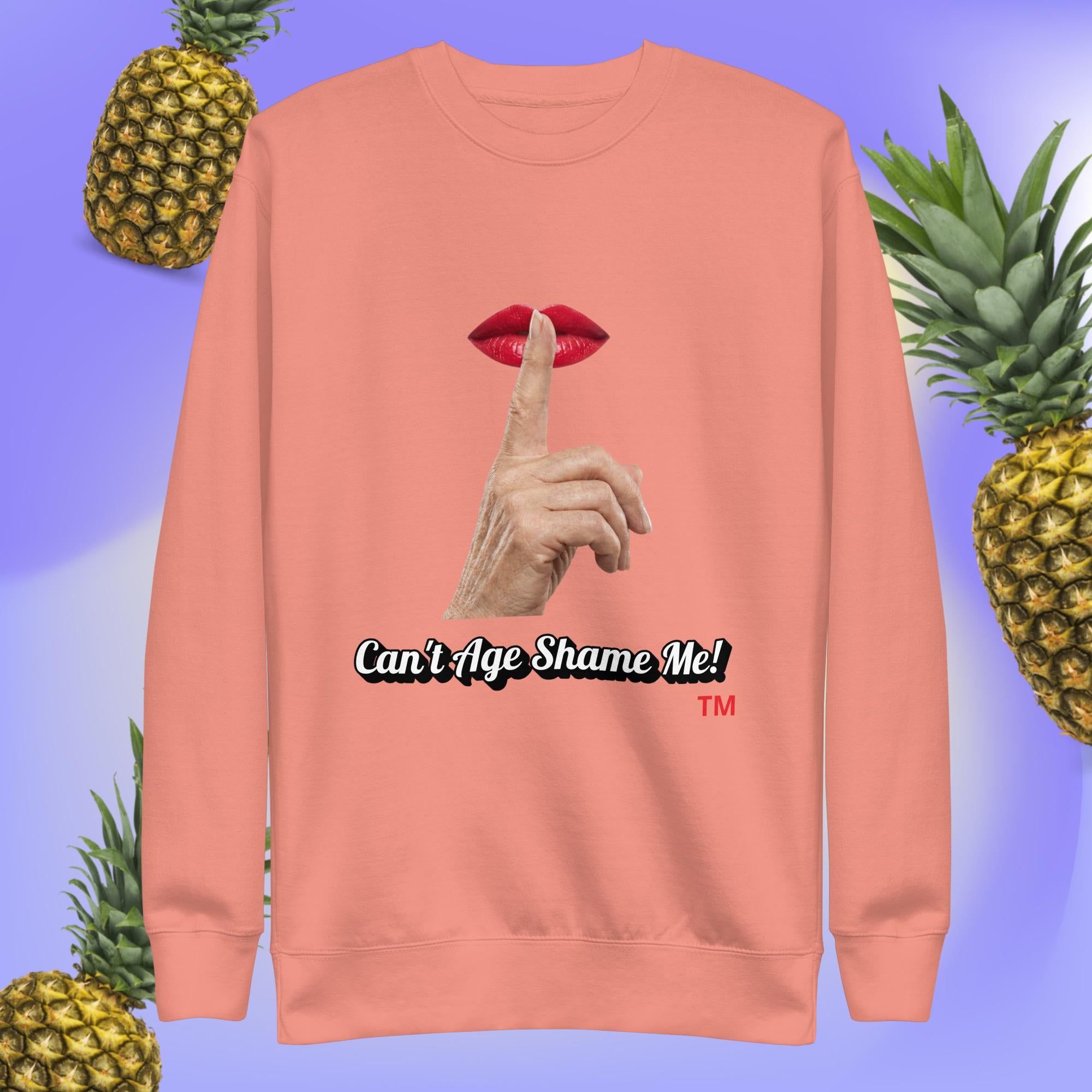 Dusty Rose sweatshirt with a graphic design of finger on mouth and text 'Can't Age Shame Me!' on a blue background with pineapples.