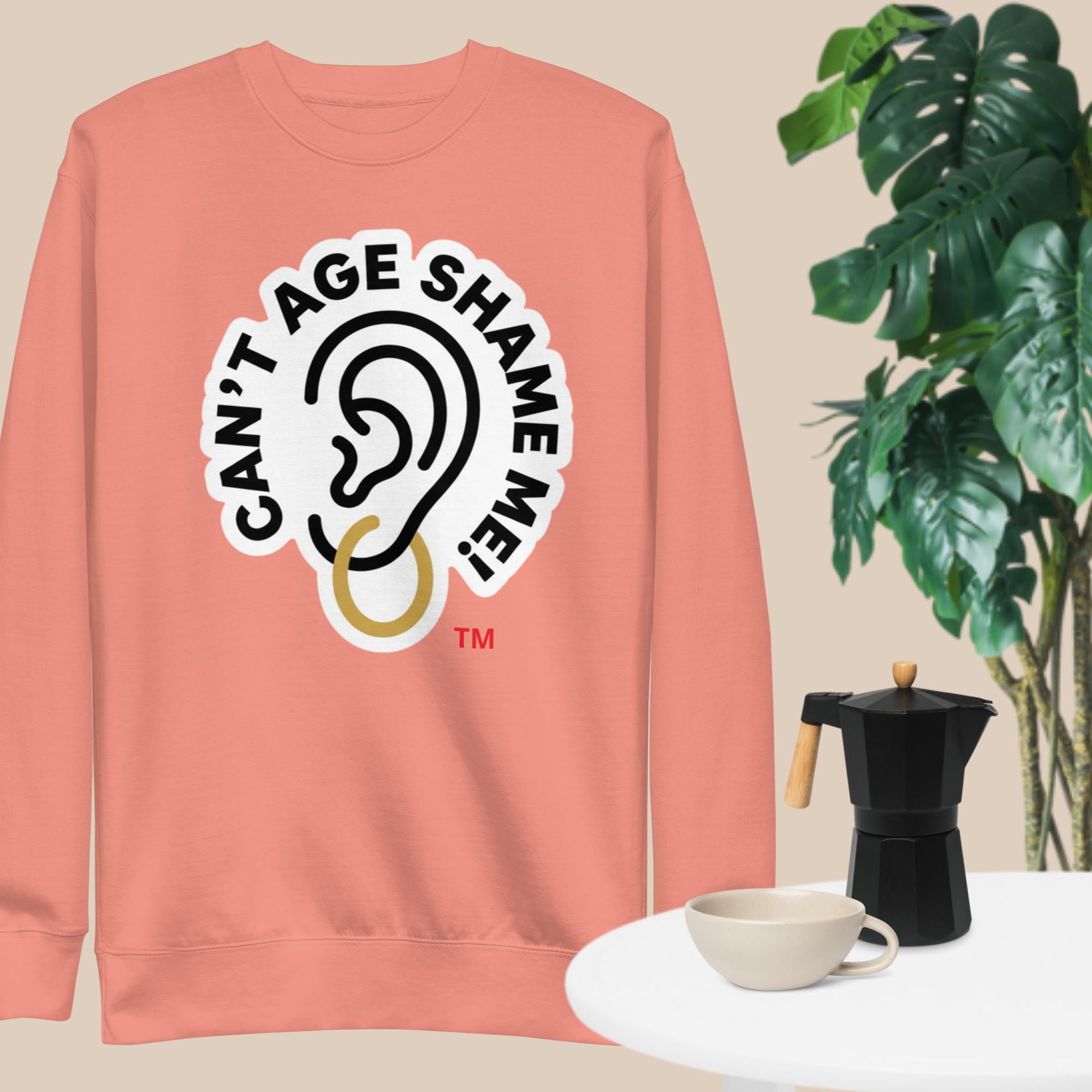 Dusty Rose sweatshirt with 'Can't Age Shame Me' graphic on a beige background with a plant and coffee set.